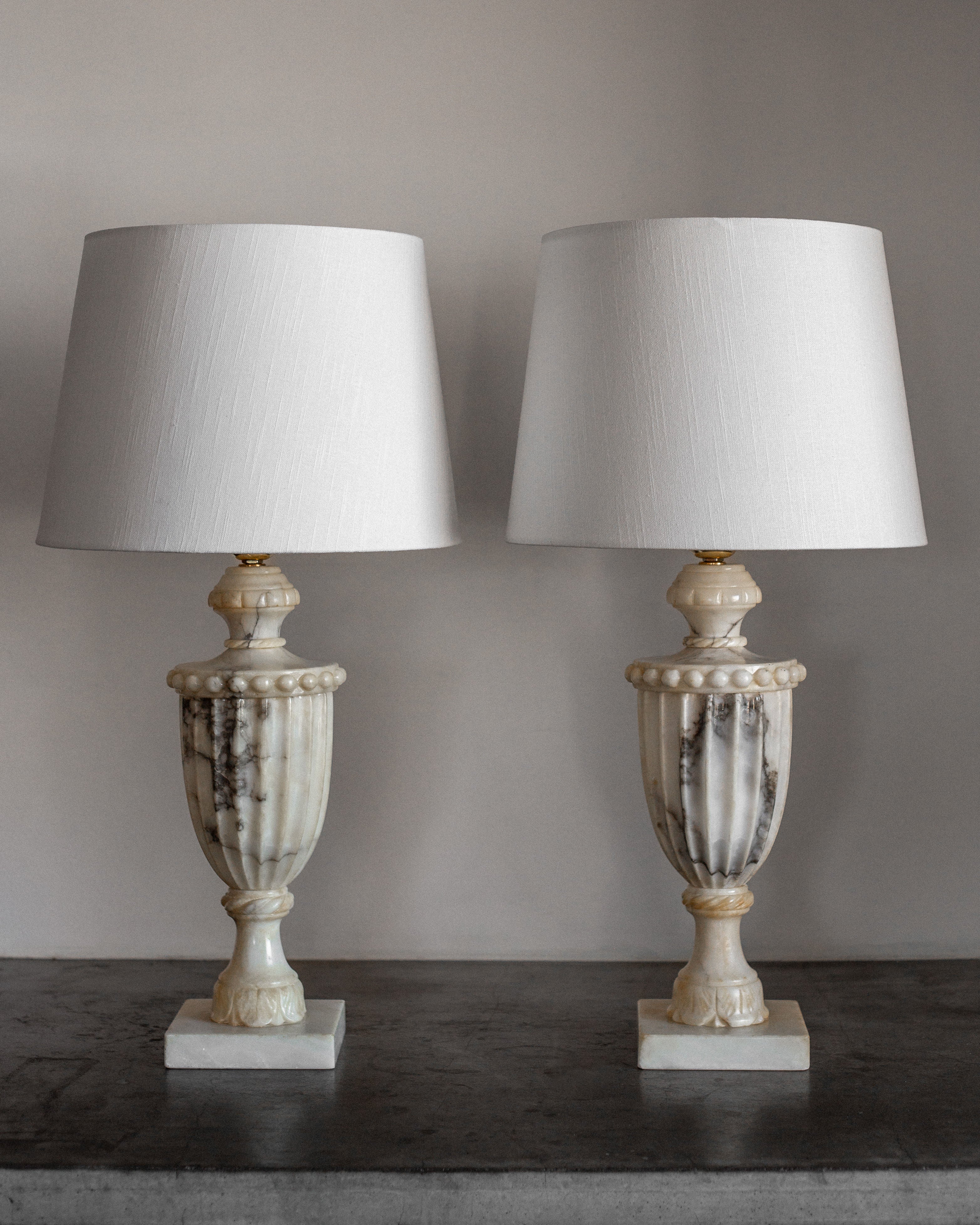 A pair of Spigel's Italian Alabaster Table Lamps, with marble-like bases and white fabric shades, stands elegantly on a dark surface against a gray wall. The urn-shaped bases showcase fine Italian craftsmanship and intricate detailing.