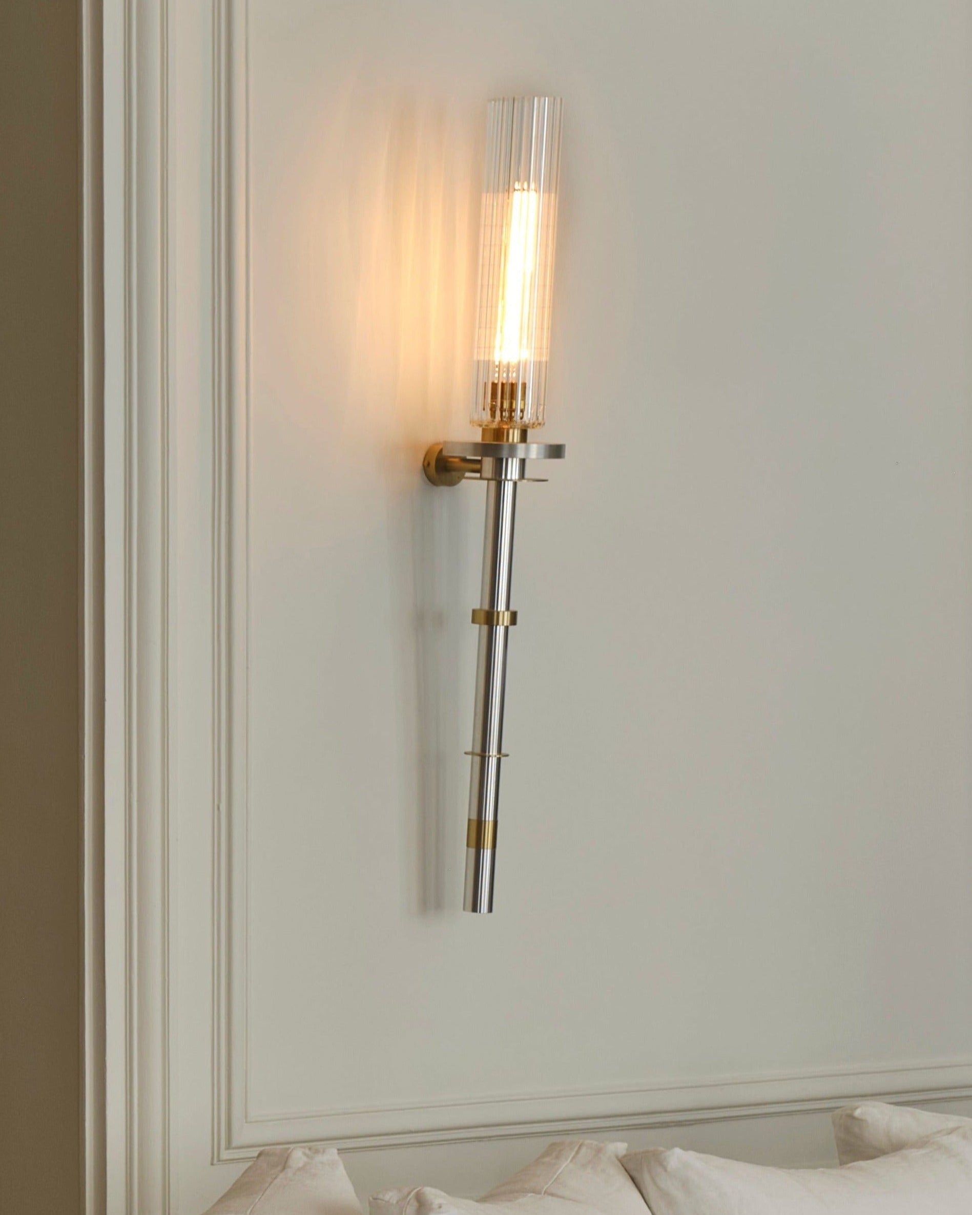 An elegant Torca Wall Light by Marine Breynaert with a striped glass lampshade and solid brass details illuminates a soft, warm glow against a pale wall above a cushioned area.