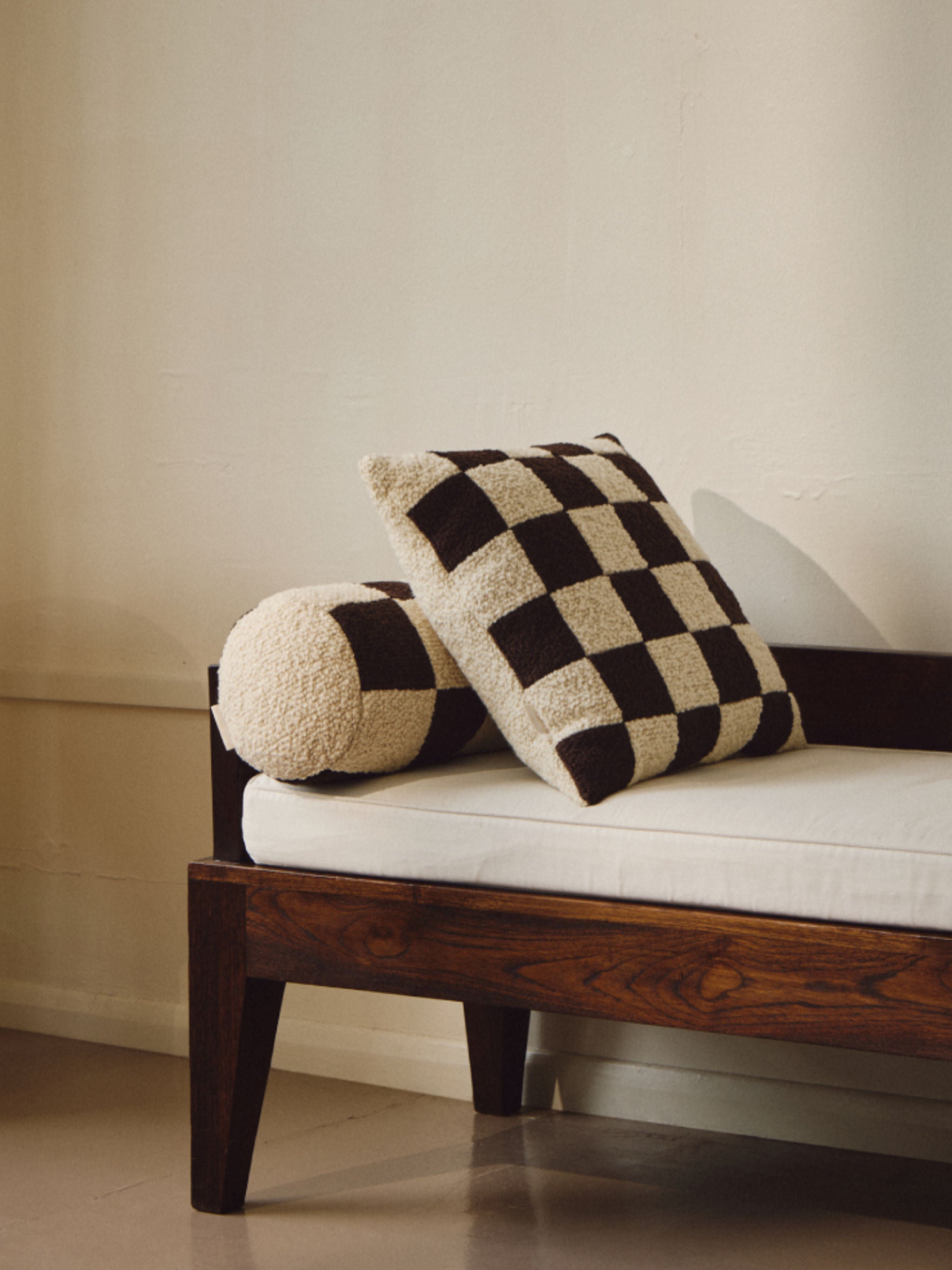 A dark wooden bench with a white cushion is adorned with two pillows: one rectangular with a black and white checkered pattern and another cylindrical one textured in cream. The Le Carré - Wool Bouclé - Chocolate/Eggshell by AMÉLIORÉ features British eco wool, while the neutral-colored wall in the background enhances the minimalist aesthetic.