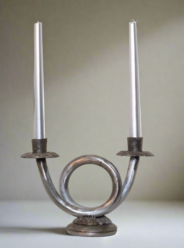 A Double Wrought Iron Candlestick by LA NOUVELLE GALERIE with a circular base and two upward, curved arms, each holding a tall, white candle. The design features a central loop, giving it a distinctive and artistic appearance. The slight oxidation adds character to its timeless beauty against the neutral background.