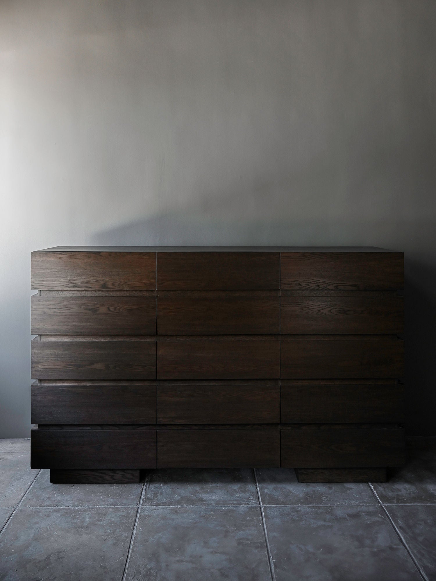 The 'Grand Château' Stained Oak Dresser by Studio OSKLO, featuring a minimalist mid-century design and nine drawers, stands against a plain gray wall on a tiled floor.
