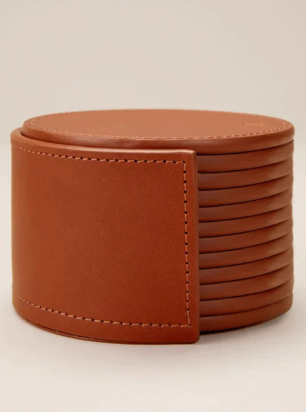 A neatly stacked set of twelve handcrafted, circular brown leather coasters from Paradise Row features visible stitching and comes in a stylish holder, showcased on a neutral background.