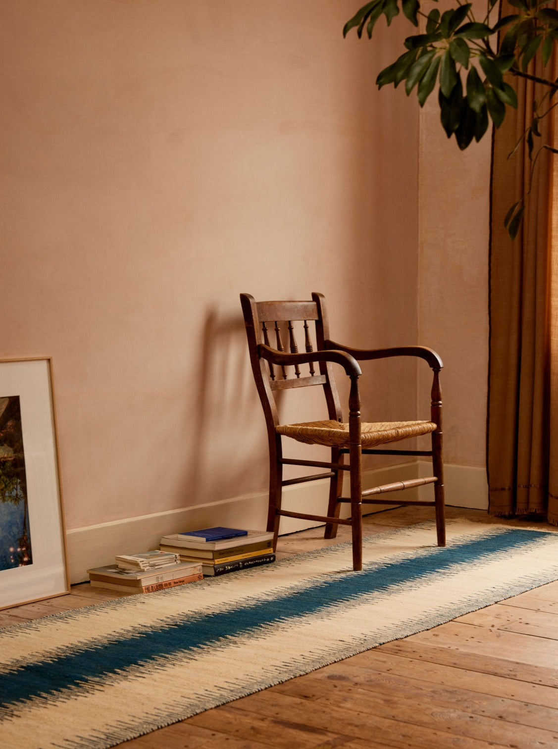 A wooden chair with a woven seat is placed against a light pink wall. Books are stacked on the floor nearby, one with a dark cover and others with lighter covers. A framed picture rests against the wall. An ISHKAR Navy Anar Kilim Runner - Handwoven Rug, patterned in natural dyes, runs along the wooden floor.