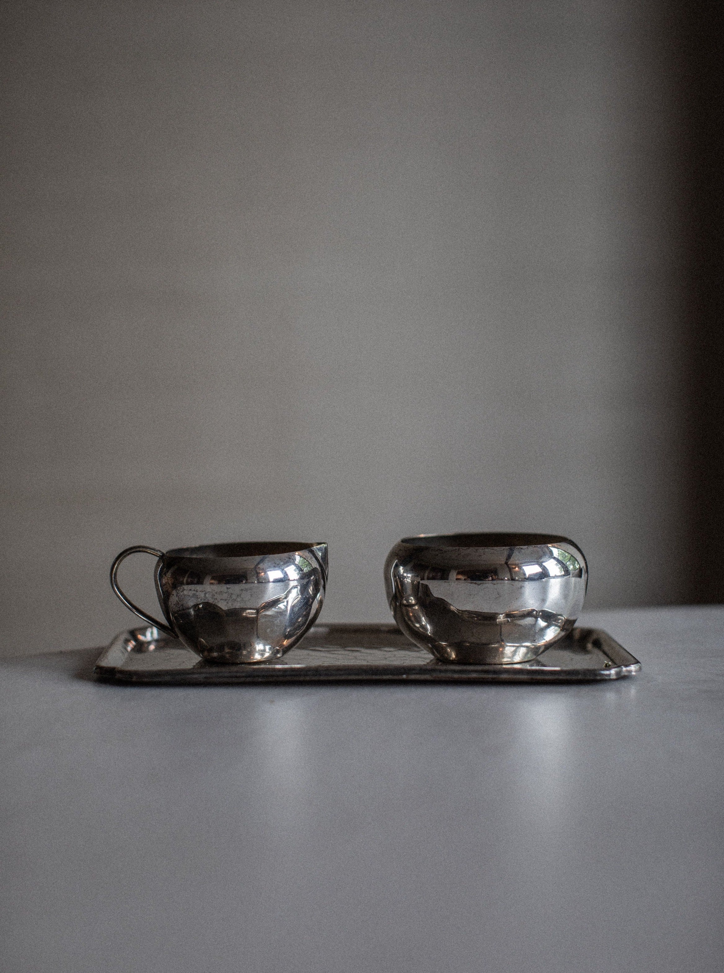 A minimalist photograph features an opulent Out For Lunch Art Deco Creamer & Sugar Bowl set placed on a matching rectangular tray. The background is softly blurred, allowing the focus to remain on the polished, reflective surfaces of the elegant tea set.