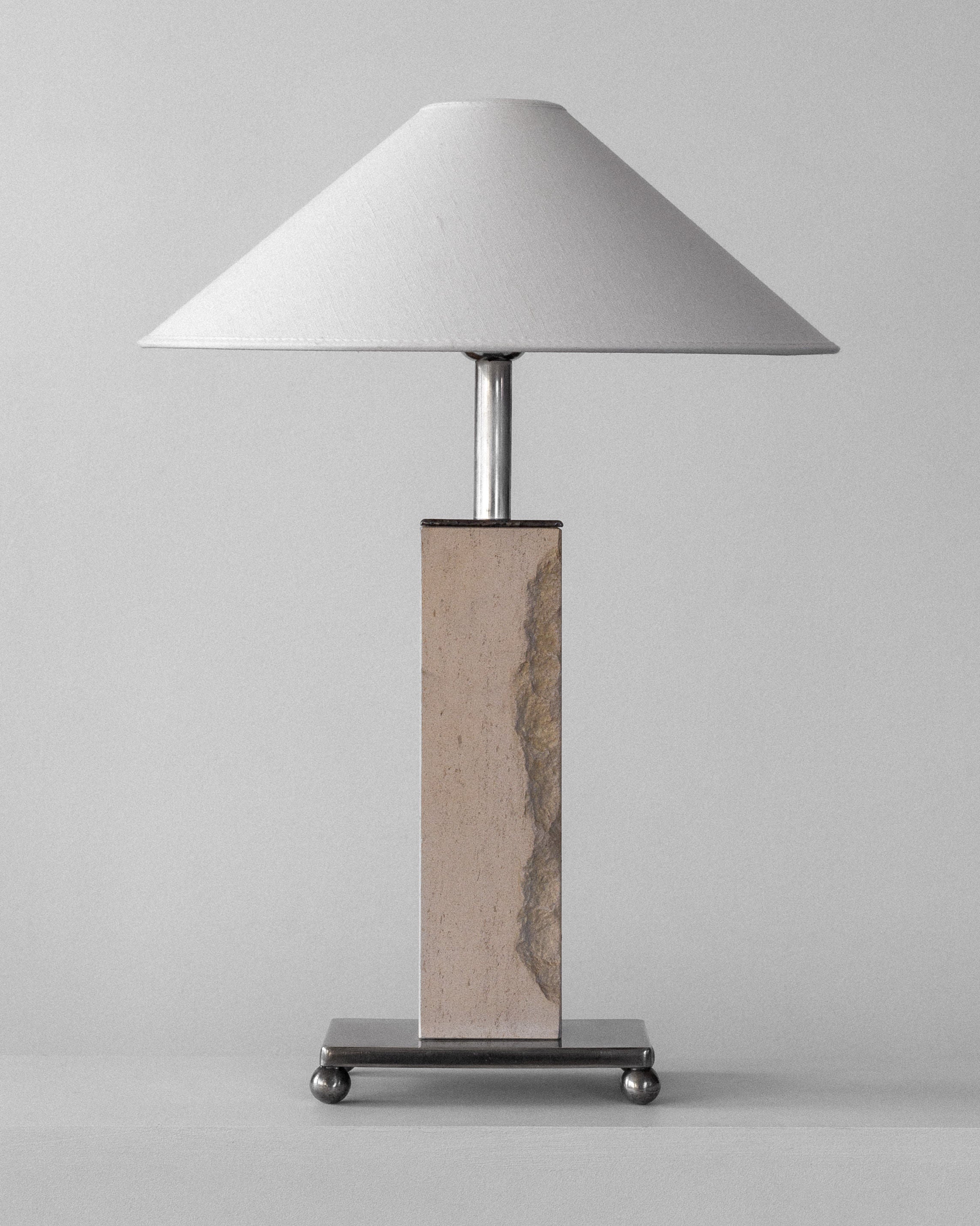 The room is graced by the Spigel Modernist Travertine and Metal Table Lamp, featuring a wide white conical shade. Its rectangular base has a stone-like finish with metallic accents, resting on a square platform with round feet. It exudes mid-century charm against a plain gray backdrop.