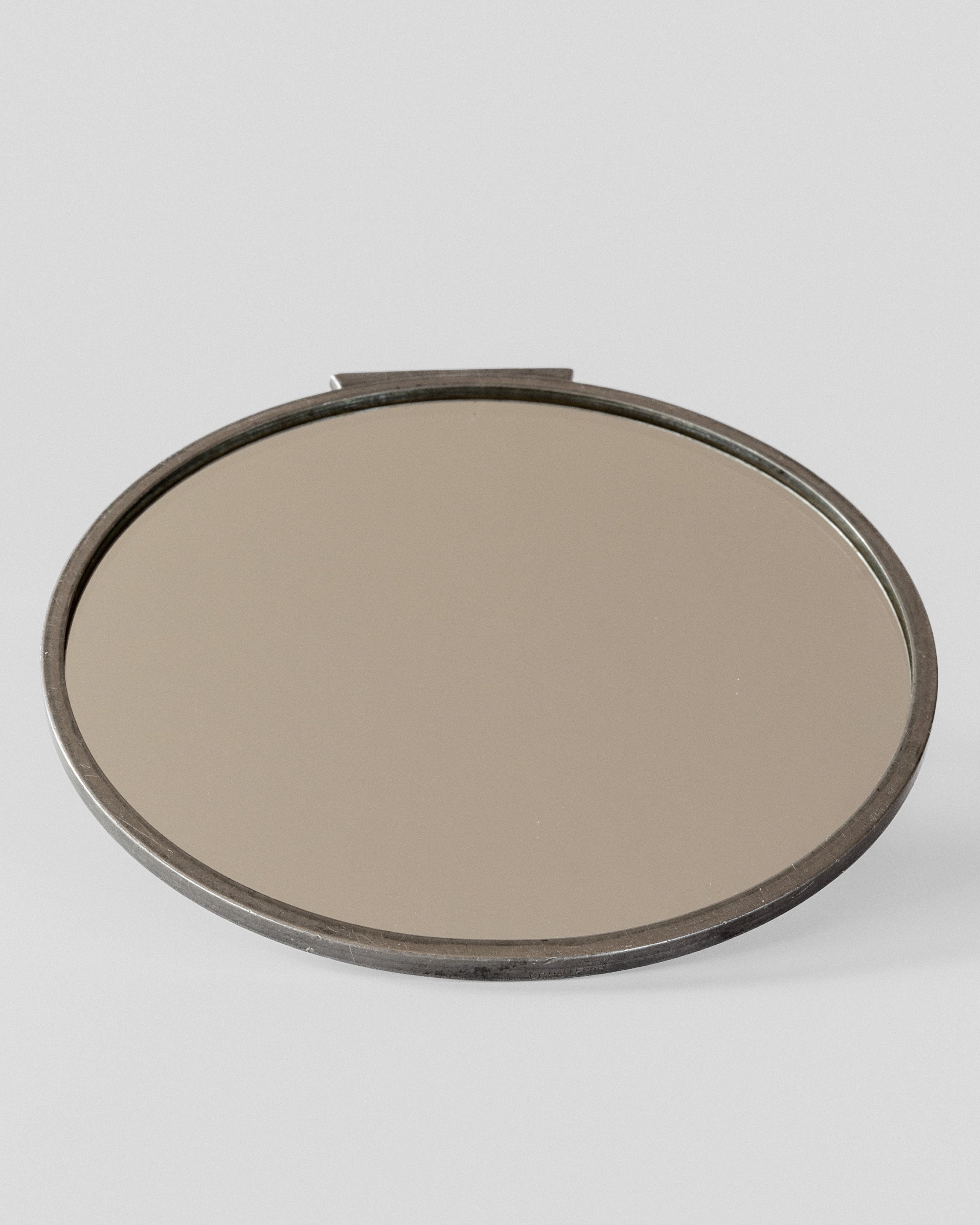 The Spigel Art Deco Metal Mirror is a round, vintage handheld mirror with a classic Art Deco style metal frame, designed to rest on flat surfaces. Its slightly tarnished appearance enhances its antique charm, highlighting its vintage condition. The silver-toned handle is partially visible, reflecting the elegance of C.G. Hallberg design.