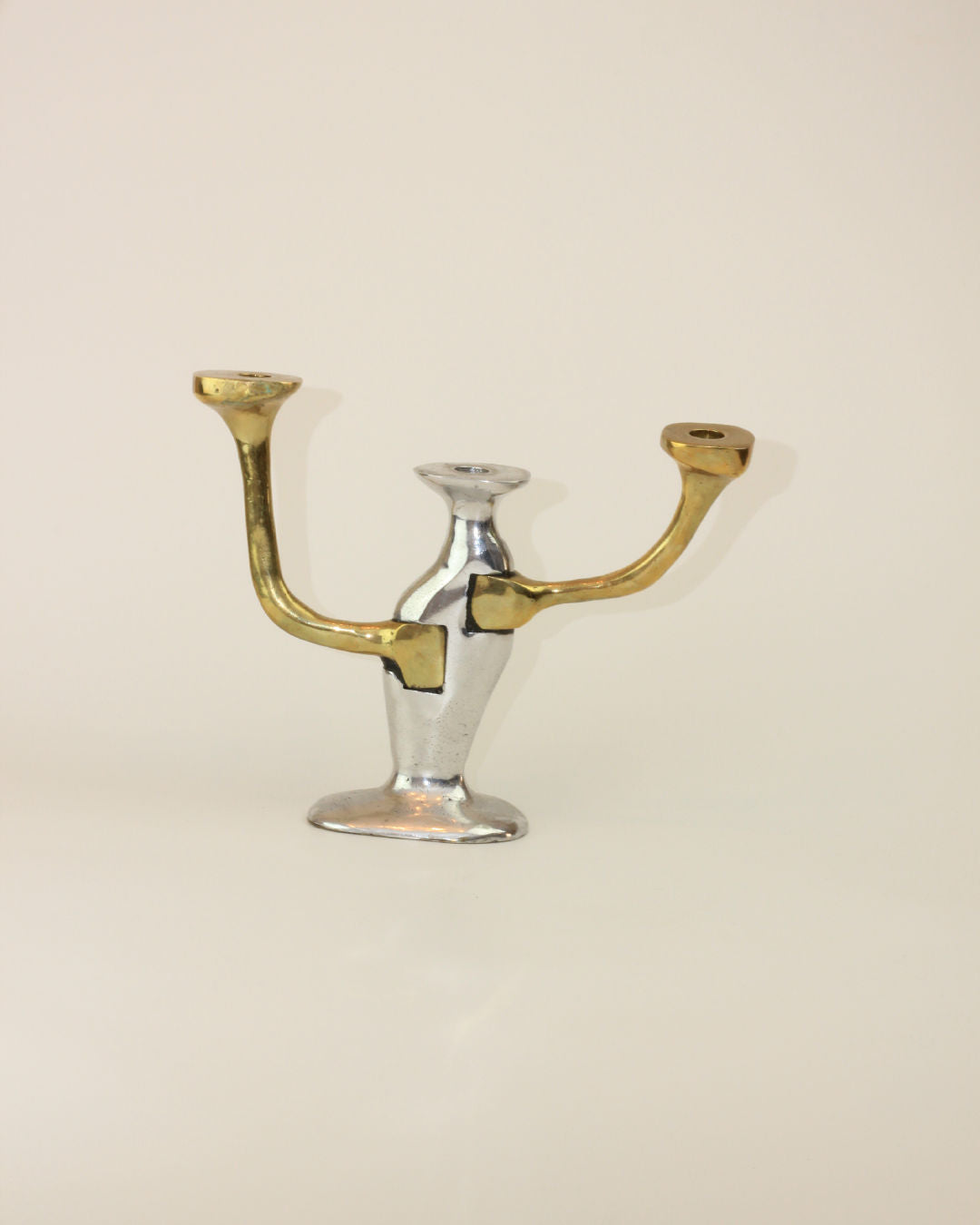 A two-armed Brutalist Candle Holder from Boga Avante Shop stands against a plain background. The base is silver, and the arms are gold, echoing a 1970s Seville design. Each arm has a depression for holding a candle. The candle holder features a simplistic and modern style with smooth curves and a sleek finish.