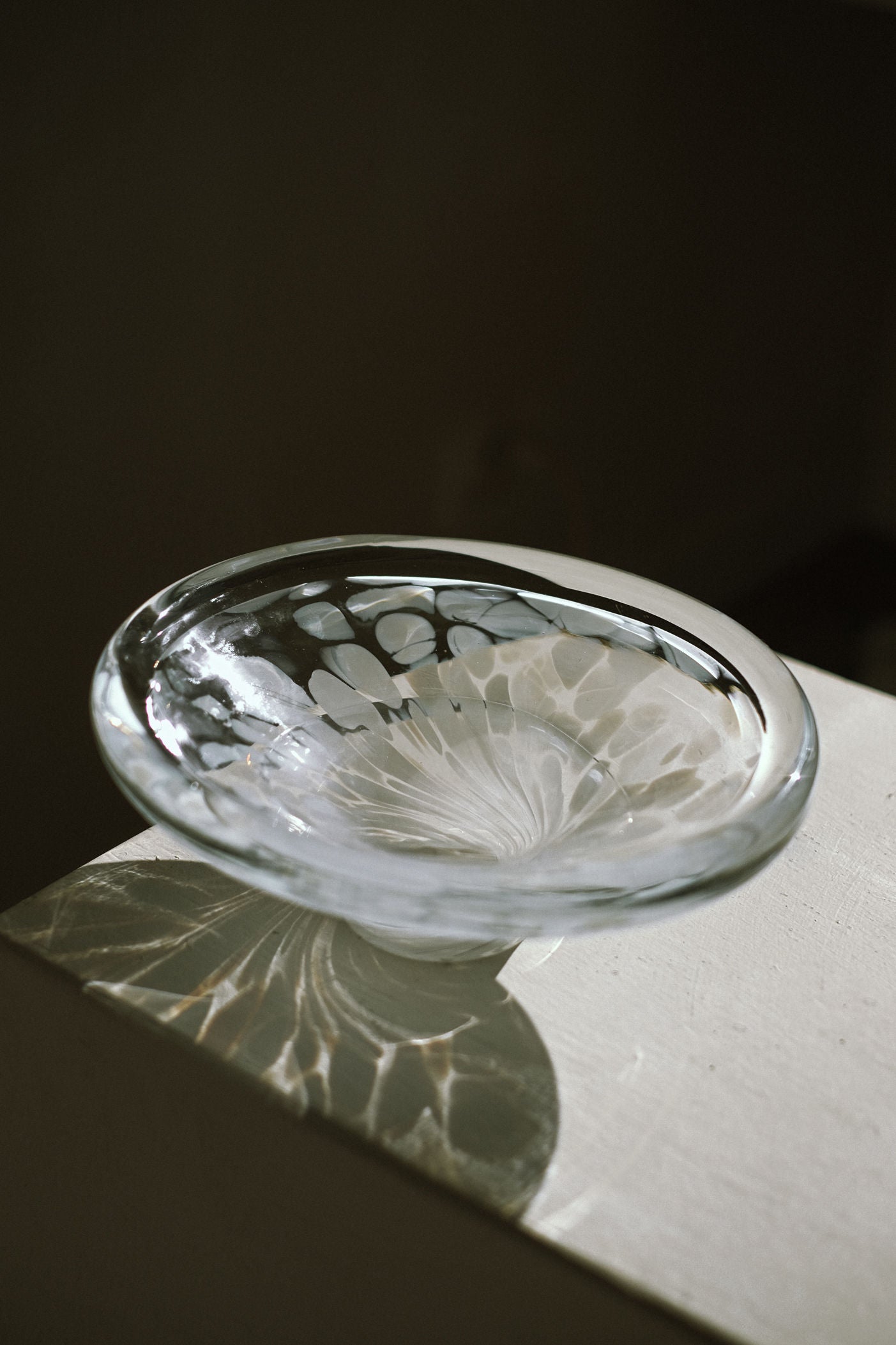 The White Bowl Tortoise Glass by septembre studios features decorative patterns and sits on a white surface, casting intricate light and shadow reflections in gentle, natural lighting.