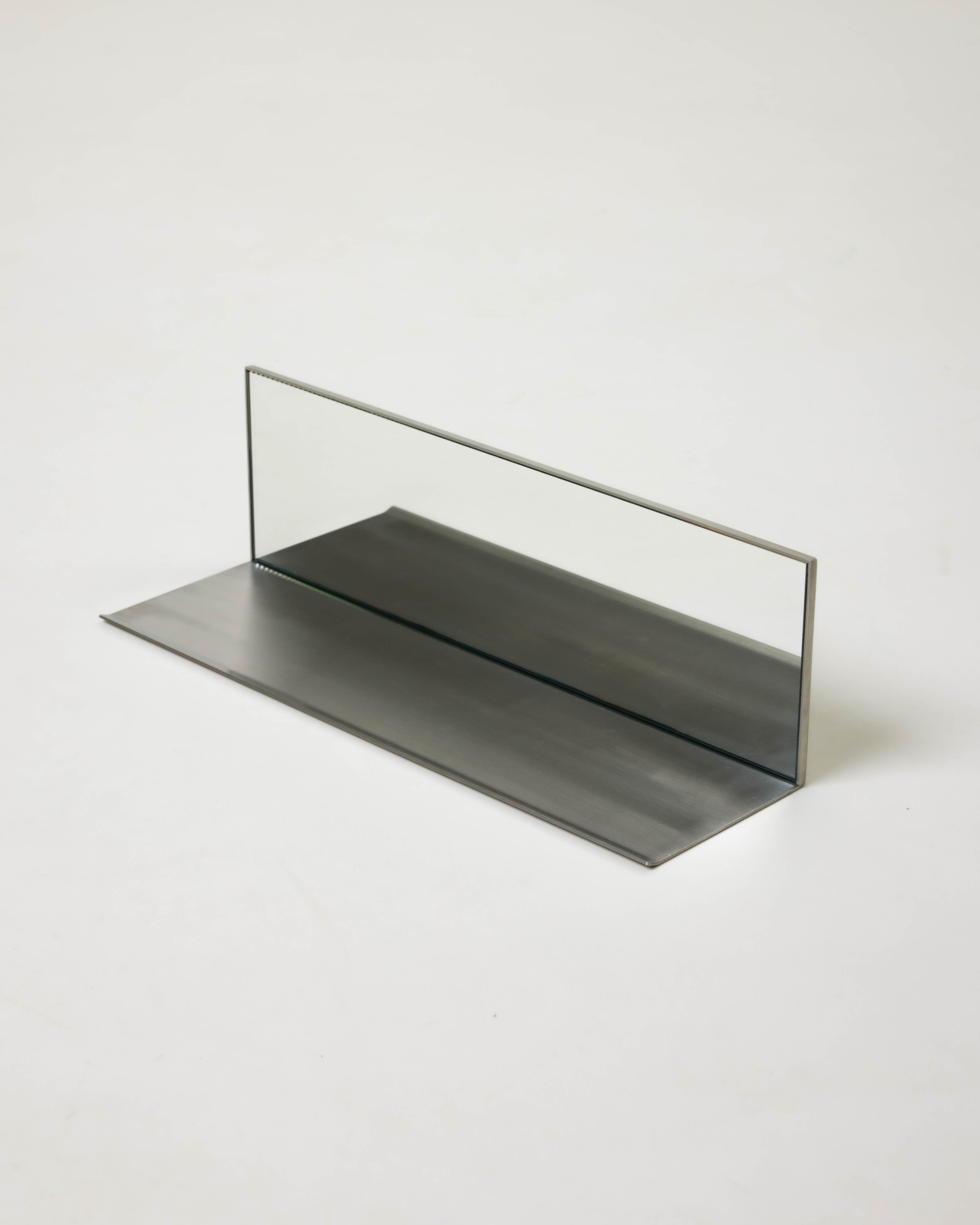 The Dot Objects Mirror Steel Shelf features a vertical rectangular glass panel standing on its reflective mirror steel base, creating a striking minimalist look against a plain white background.