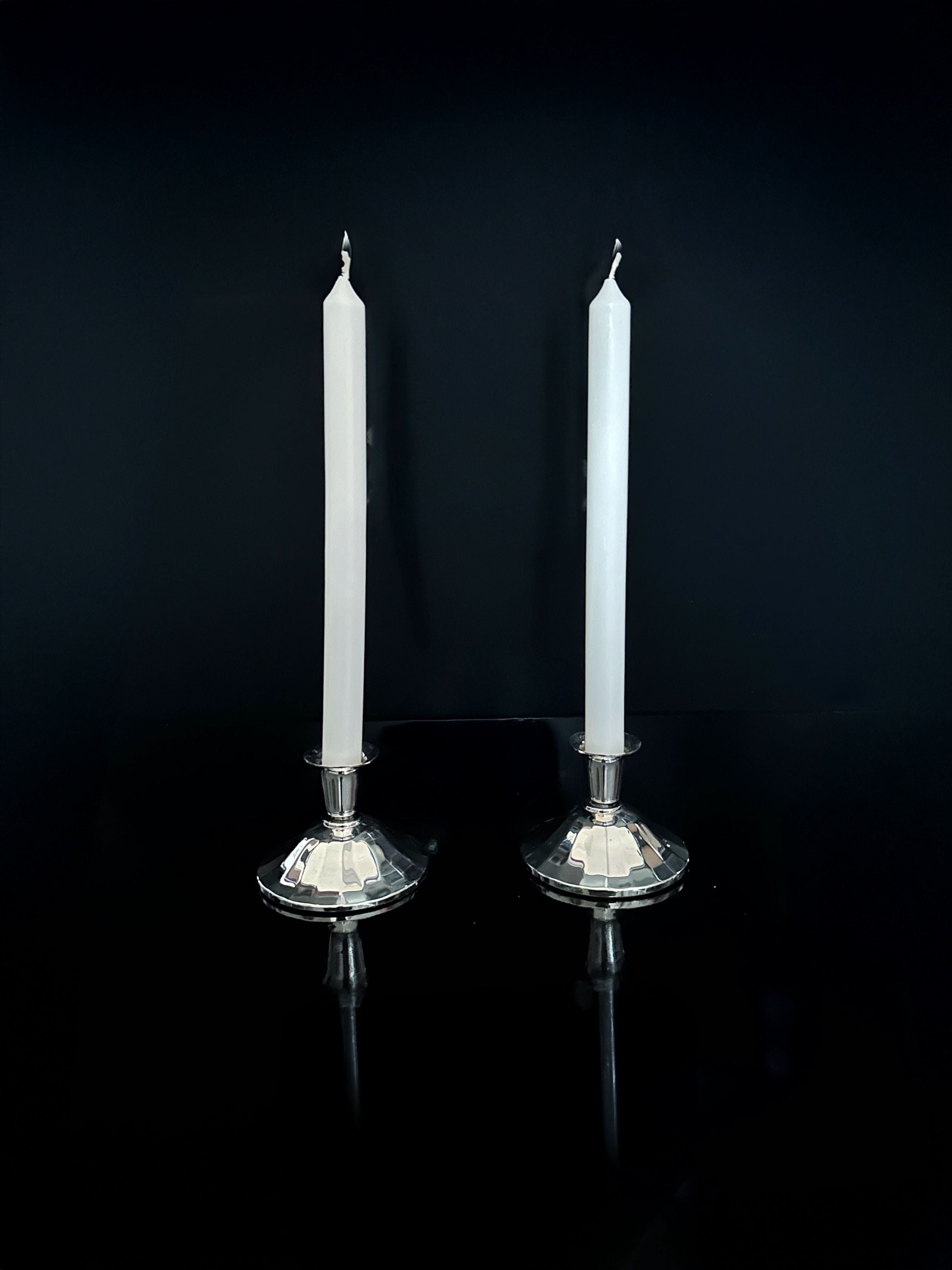 Two tall white candles are positioned in Silver-Plated Art Deco Candleholders from 1930 by Dodo Vintage, standing side by side against a dark backdrop. The candleholders and candles are beautifully reflected on the glossy surface below.
