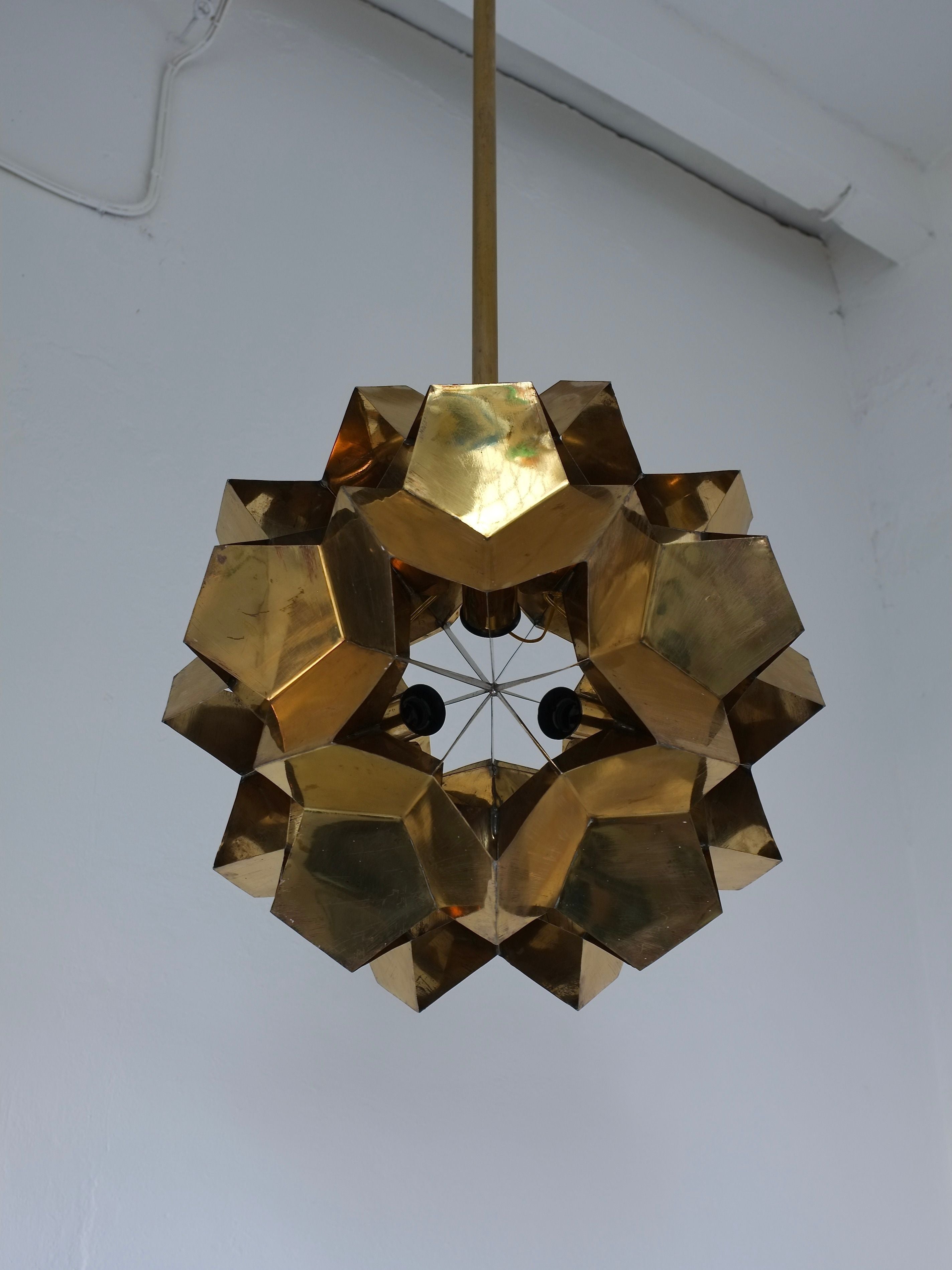 The Brutalist Brass Chandelier by Veter Vintage features a geometric dodecahedron design in solid brass, inspired by 1970s Latvia. This sculptural ceiling pendant is E27 bulb compatible and stands out against a white wall.