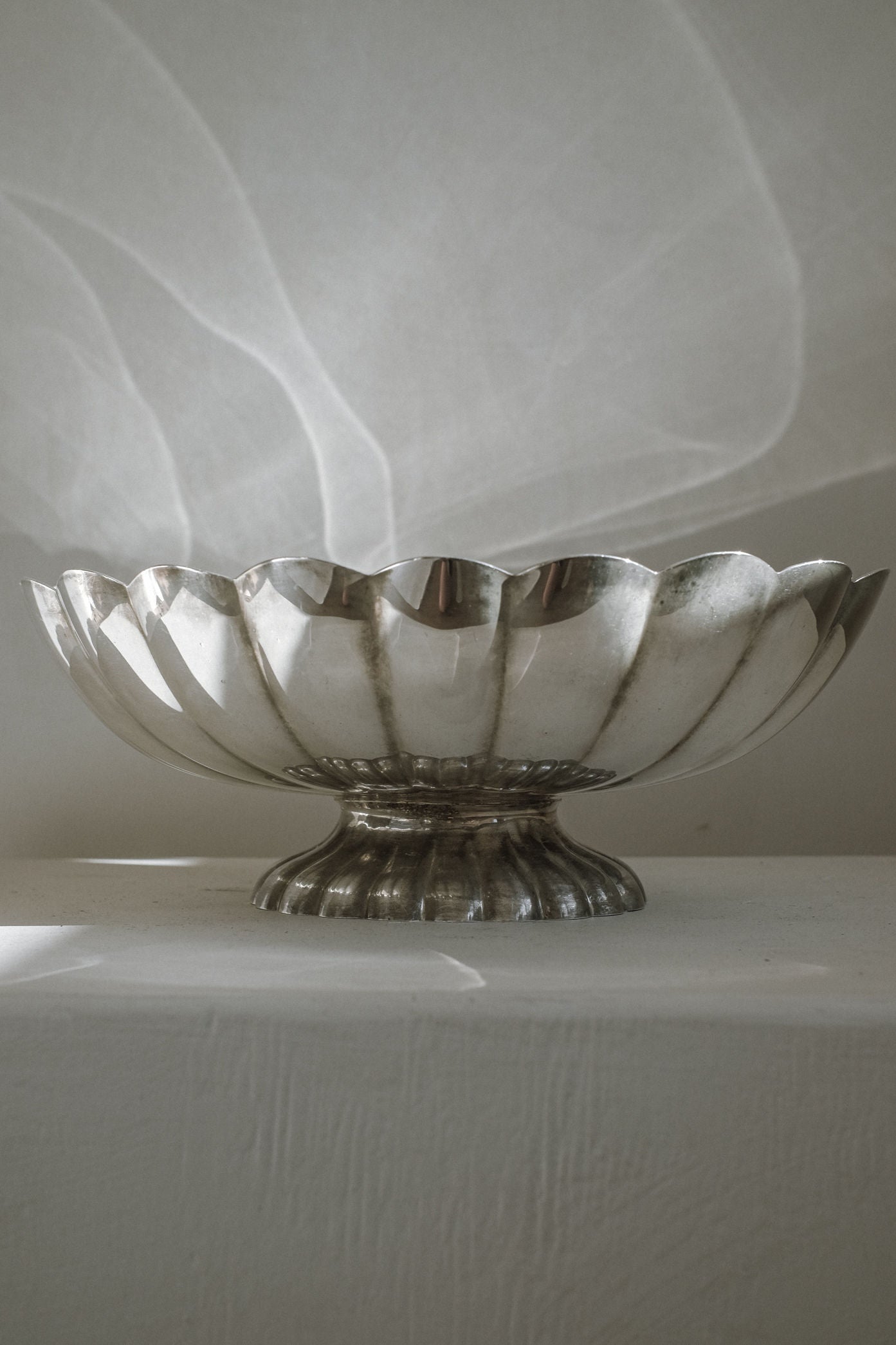 A Silverplated Centerpiece by septembre studios with scalloped edges and a short pedestal base rests on a white textured surface, softly lit to reveal gentle shadows and reflections against a plain background.