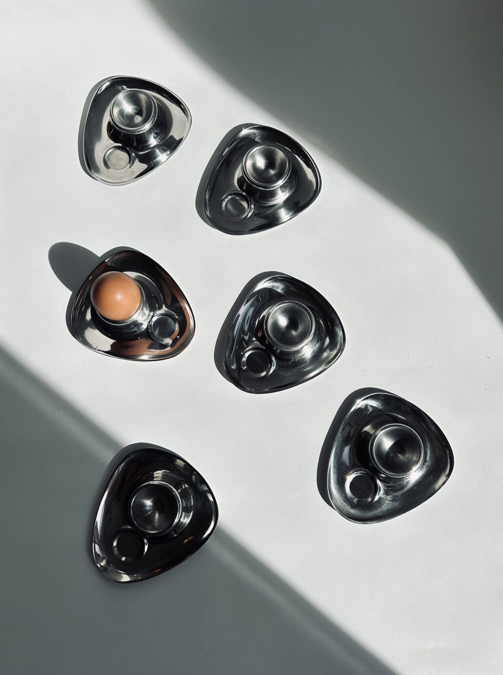 A set of six Bauhaus Stainless Steel Egg Cups from Flock Studio is displayed on a white surface, each featuring two circular sections in a triangular design. One of these sleek holders cradles a brown egg, capturing the essence of vintage mid-century charm. Shadows across the image add to its dramatic allure.