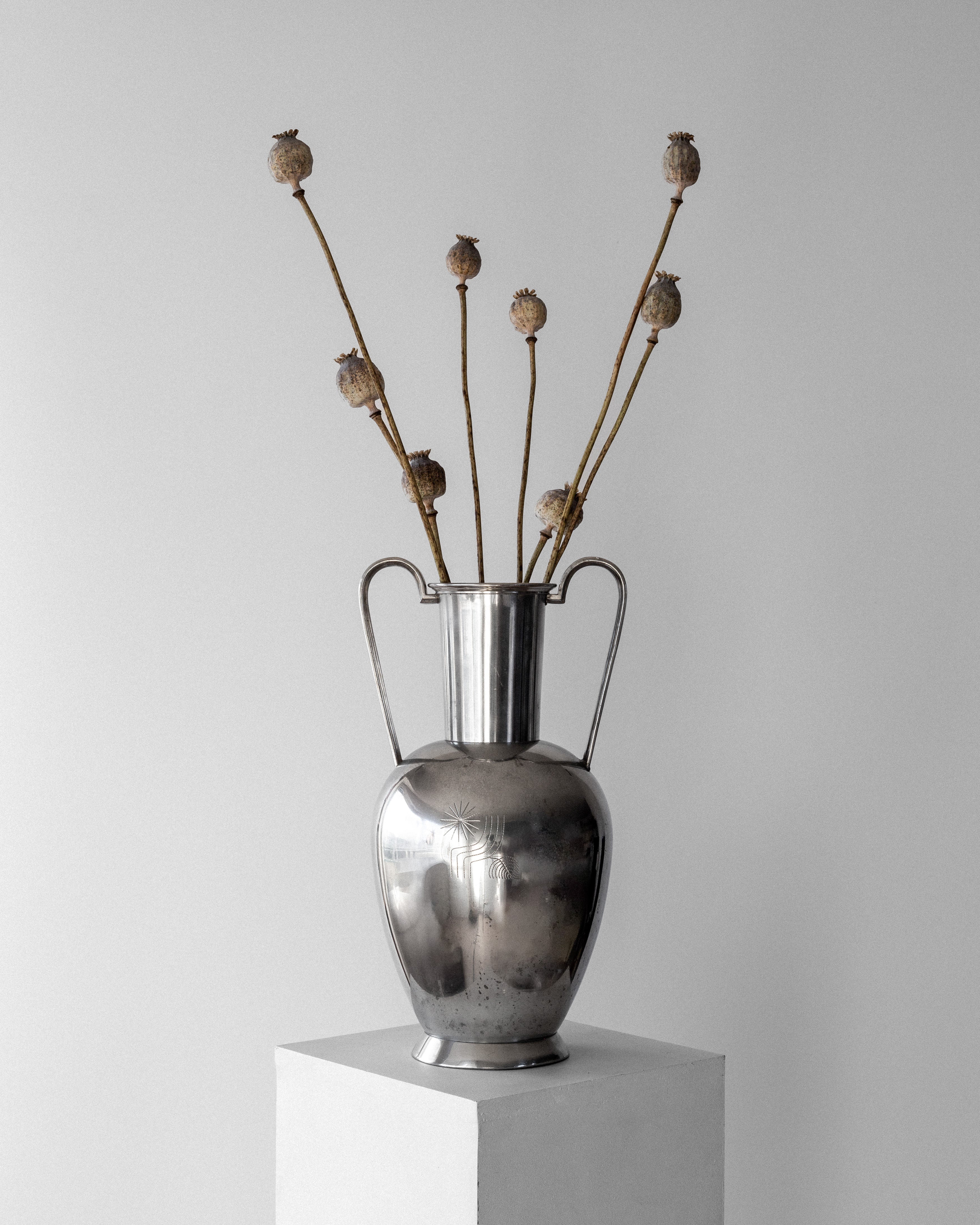 A Large Modernist Pewter Amphora by Spigel, reminiscent of Scandinavian metalwork from the 1930s, with double handles holding dried poppy stems, is elegantly placed on a white pedestal against a plain light gray background.
