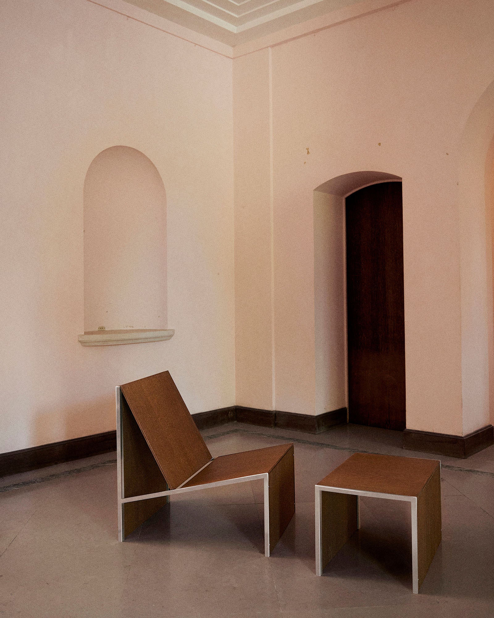 A minimalist room with pale pink walls showcases Studio HAOS’s Lounge Chair 2022 and matching low table in wood, placed on a light gray floor near arched doorways and a wall niche, embodying grounded elegance.