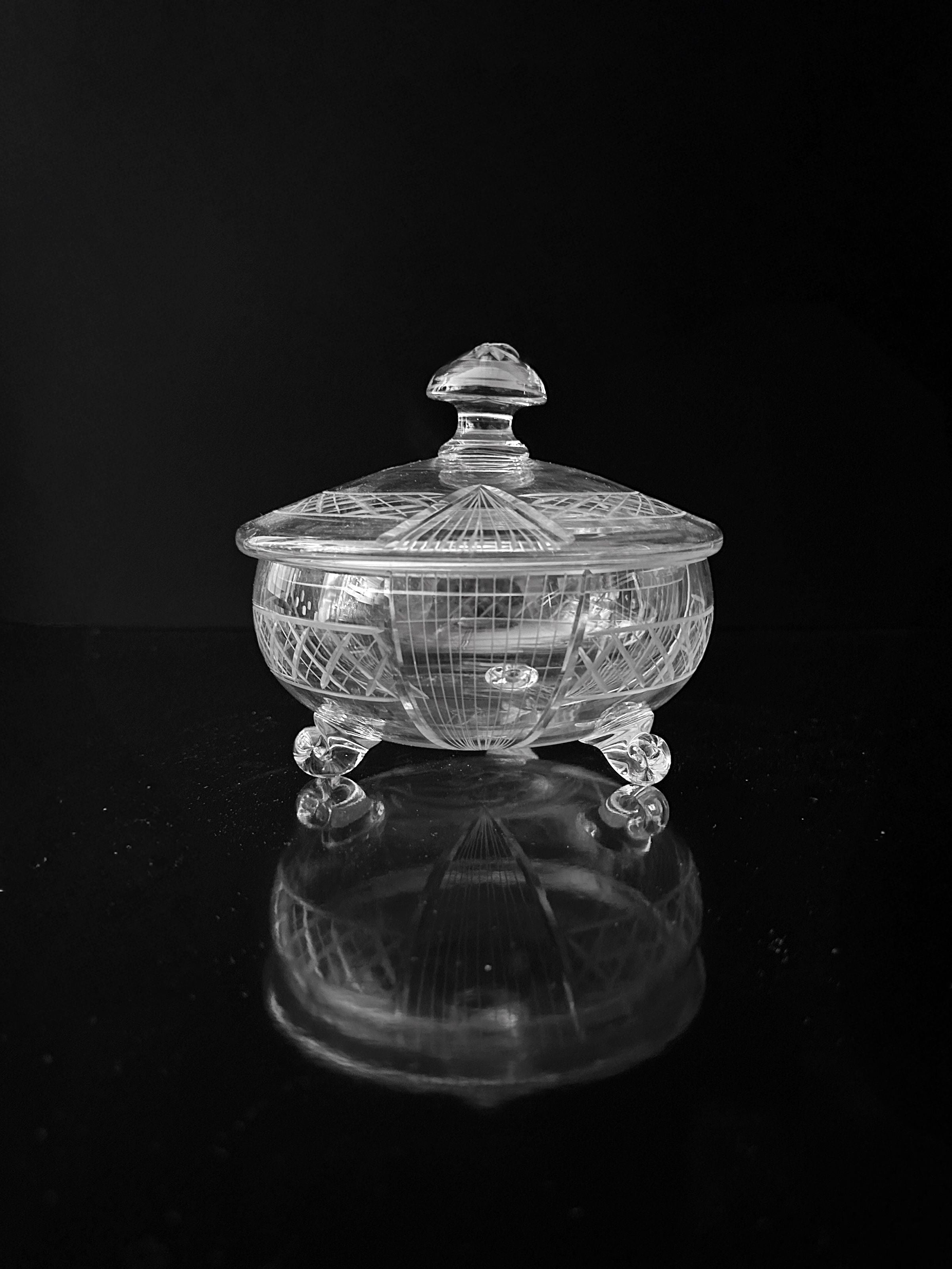 The Dodo Vintage Art Deco Glass Bonbonnière - 1930, a handmade piece with a lid, sits elegantly on a reflective black surface. It showcases intricately engraved geometric patterns reminiscent of the Art Deco era and is gracefully supported by three small decorative legs.