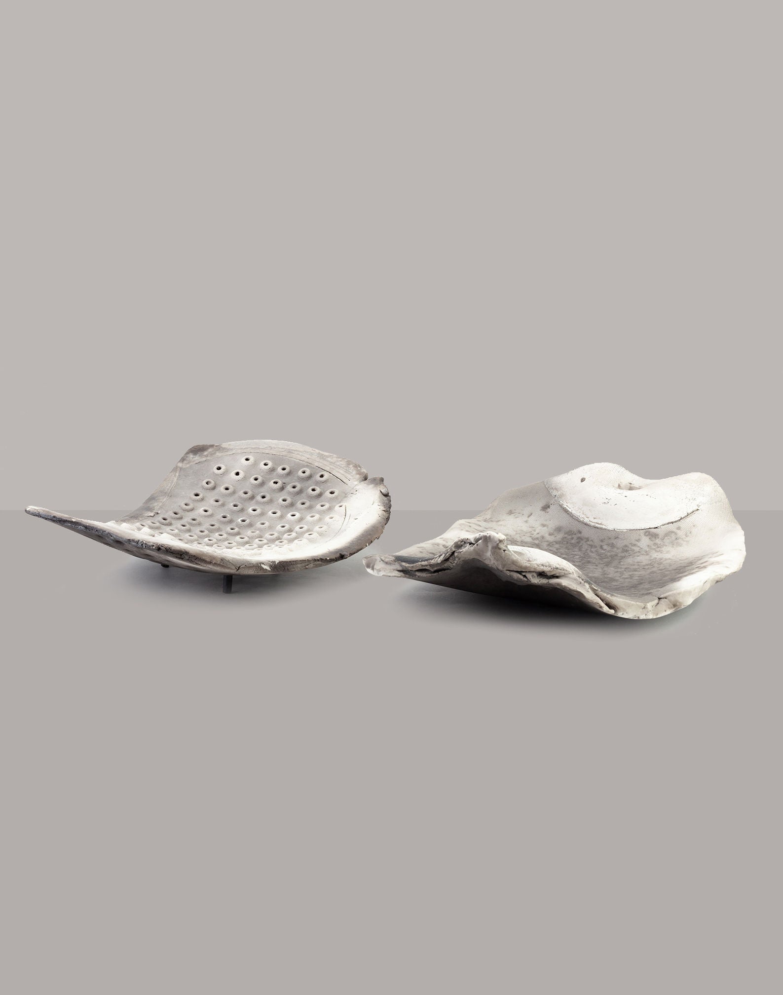 Two freeform Raku platters by John Dunn, from RELIC LONDON, showcase an abstract leaf-shaped design in shades of gray. The left piece features a pattern of small holes reminiscent of a ceramic sculpture, while the right piece displays a smoother surface and rests gracefully on tripod feet against a neutral gray background.