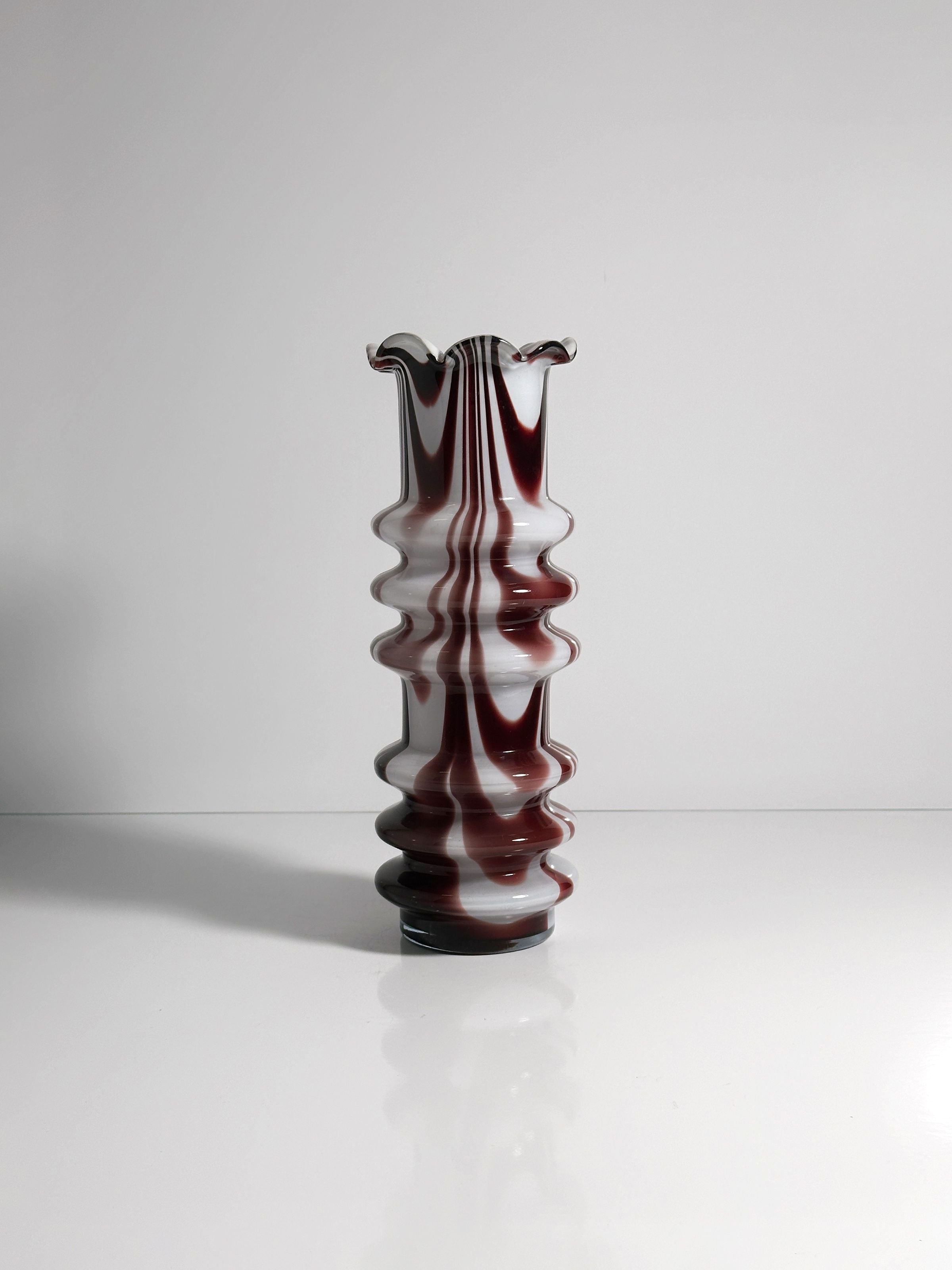 A 1970s Carlo Moretti Opalina Fiorentina Vase by Dodo Vintage, featuring a wavy design with red and white marbling, rests on a reflective white surface against a plain backdrop. The vase has a ruffled top and vertical ridges, casting a soft shadow.