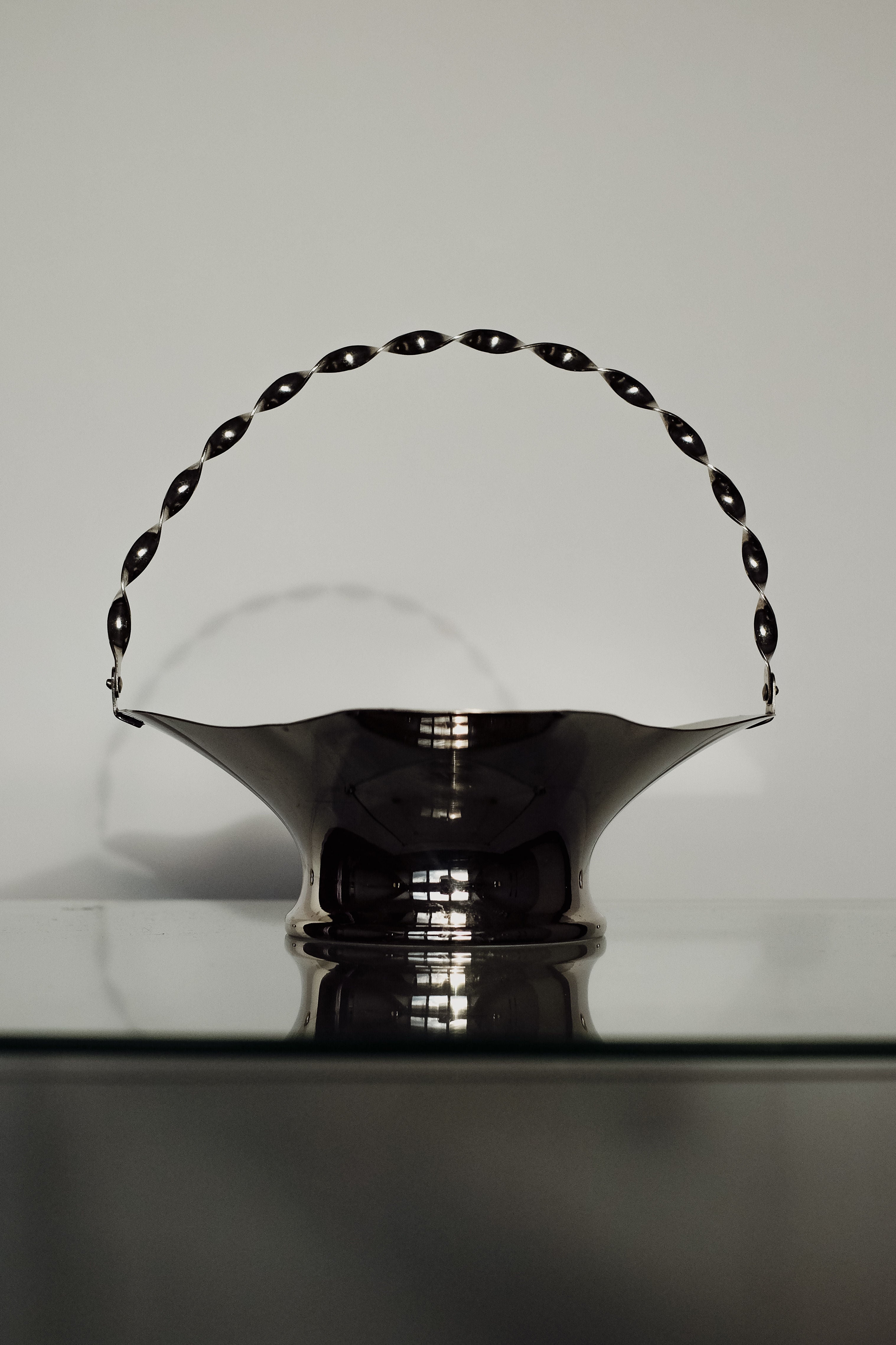 The "Basket With Twisted Hand Barrel" by septembre studios showcases unique character with its sleek black metal design and decorative handle. It rests gracefully on a glass surface against a light gray background, featuring smooth curves and an elegant, minimalist aesthetic.