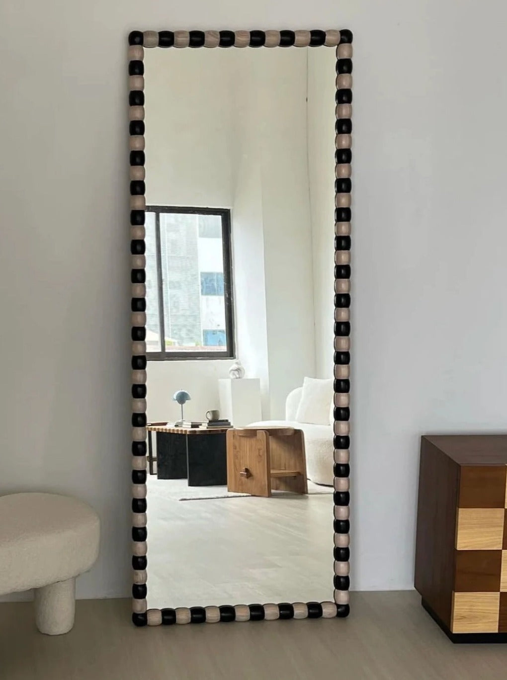 A rectangular, full-length mirror with a handcrafted solid wood frame is propped against a wall. The mirror, the Onde Mirror (Large) by Studio Kallang, reflects a modern, minimalistic living room that includes a black coffee table, a wooden side table, a white sofa, and tall windows.