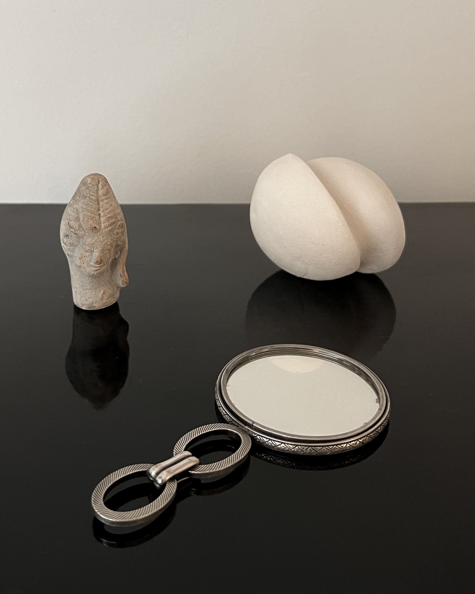 A small stone face sculpture, an oval white sculpture on its side, and the Collection apart's Hand Mirror Jean Després 1920s with a beautifully ornate double-looped handle are elegantly displayed on a glossy black surface.