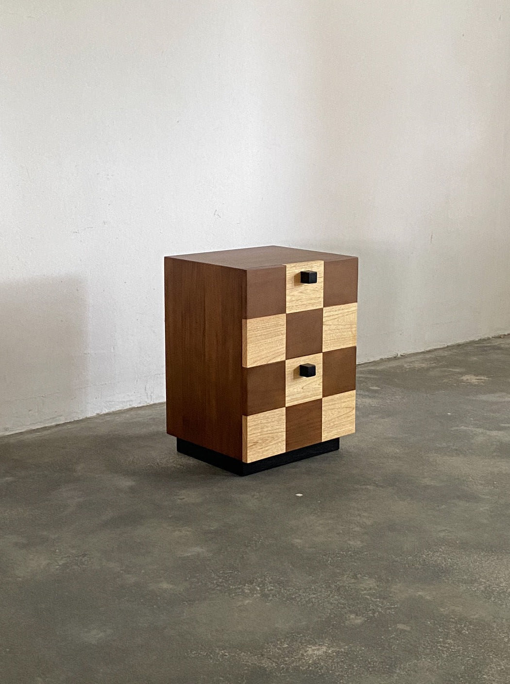 A wooden storage cabinet stands against a plain, off-white wall, showcasing a checkerboard facade with alternating light and dark wood panels. The Coklat Night Stand by Studio Kallang includes three storage drawers, each adorned with square black handles, and stands on a sleek black base.