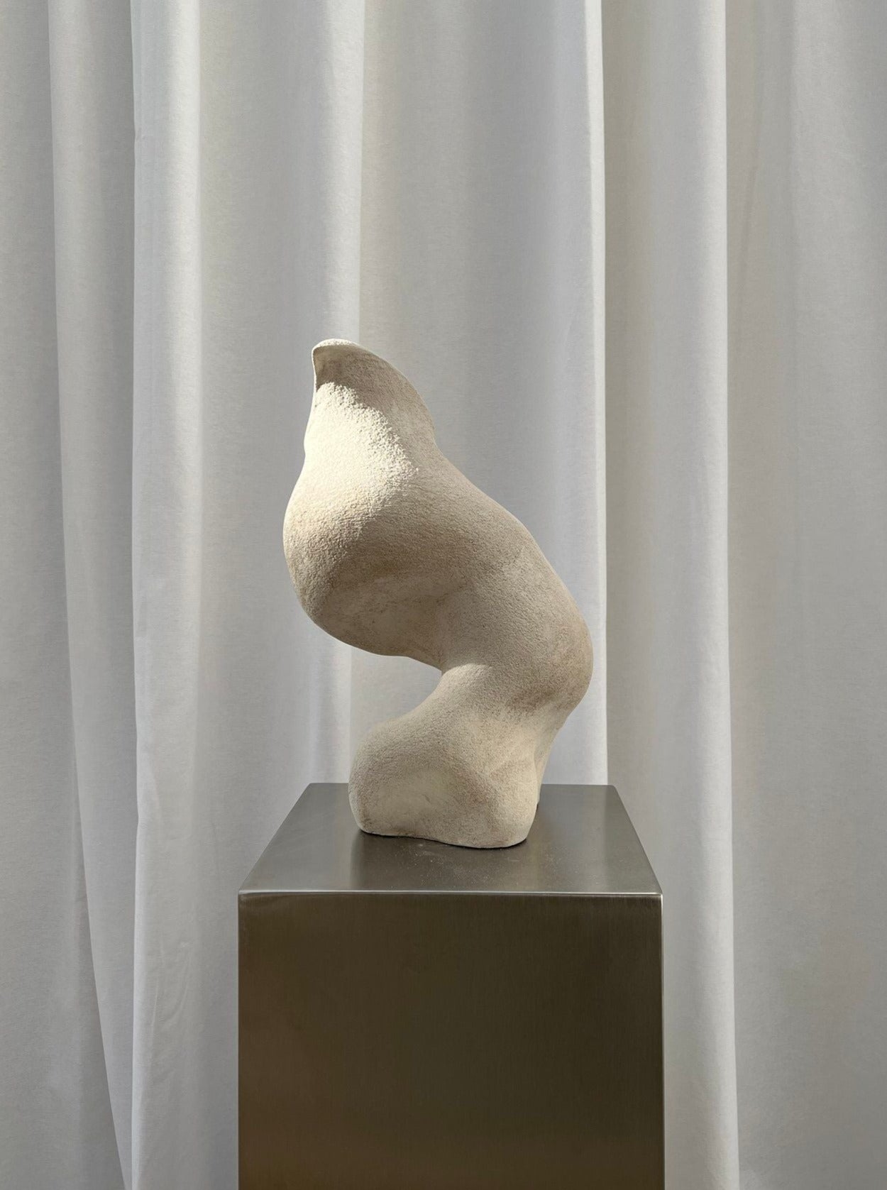 A Sculpture Seashell of a partial abstract human figure, focused on a thigh and torso, crafted from high-fired stoneware and displayed on a metallic pedestal with a draped white curtain background by 2222Studio.