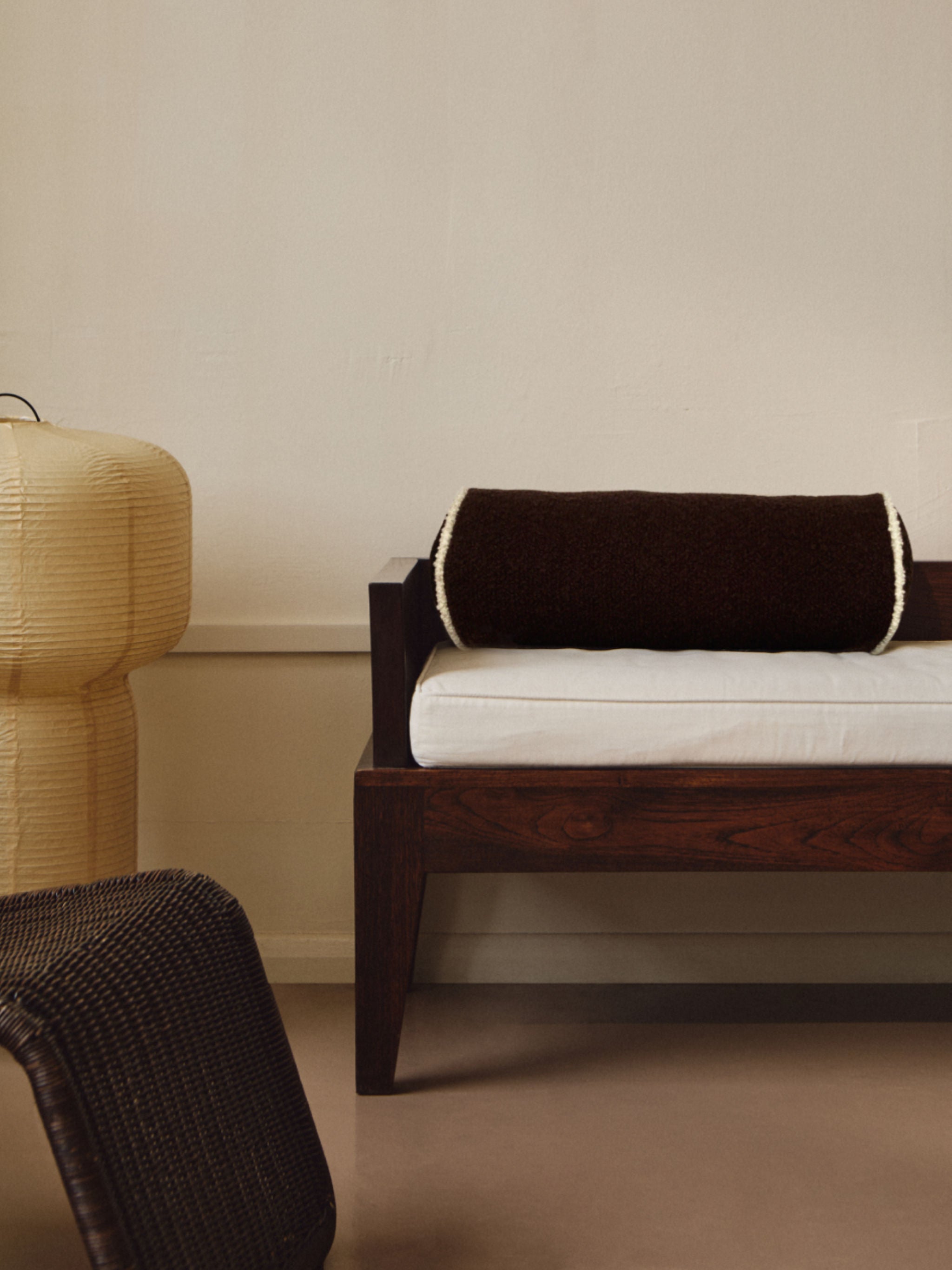 A minimalist room featuring a wooden bench with a white cushion and AMÉLIORÉ's Le Cylindre - Passepoil - Wool Bouclé - Chocolate/Egg White. To the left is a beige, textured lamp, and in the foreground is a wicker chair. The wall and floor are neutral tones, creating a serene atmosphere.