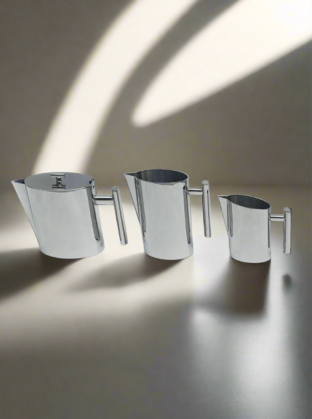 Three sleek, stainless steel pitchers of varying sizes are lined up against a neutral background. A beam of sunlight casts a soft shadow, adding a minimalist and modern aesthetic to the Les Objoies Vintage Coffee Set composition.