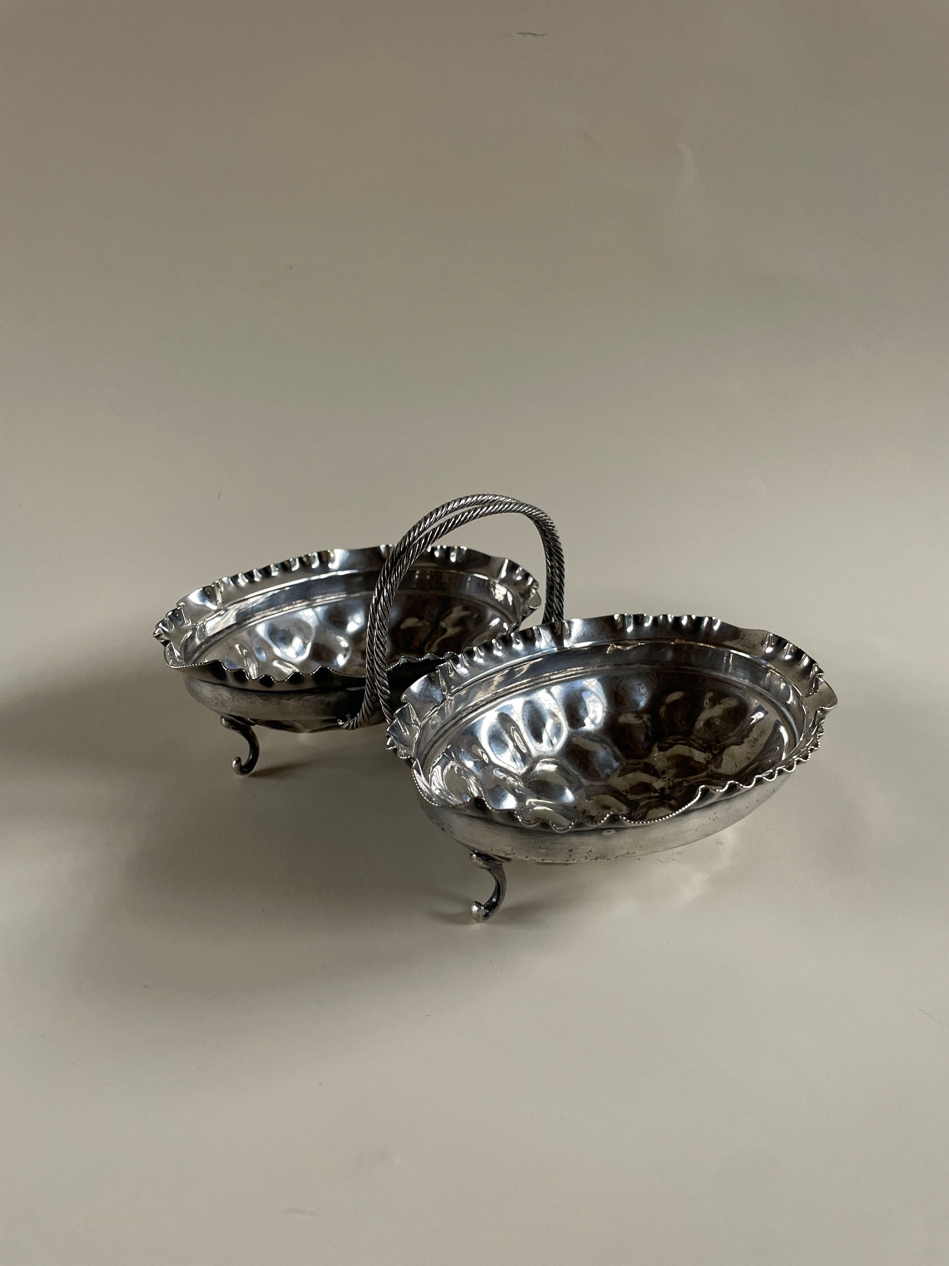 Presenting the Maud Vaughan Silver Plate Serving Dish, crafted with two interconnected oval bowls and a central decorative handle. Its scalloped edges add a touch of charm, while three small legs provide an elegant stance, all accentuated by a reflective finish. A plain and neutral background enhances its sophisticated allure.