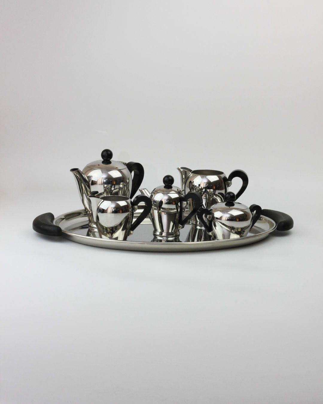 The "Bombè" Alessi Tea/Coffee Set from Boga Avante Shop, exemplifying mid-20th-century craftsmanship in silver, boasts a tray with black handles and polished teapots, creamers, and a sugar bowl—each adorned with black knobs and handles—neatly arranged on a white surface.