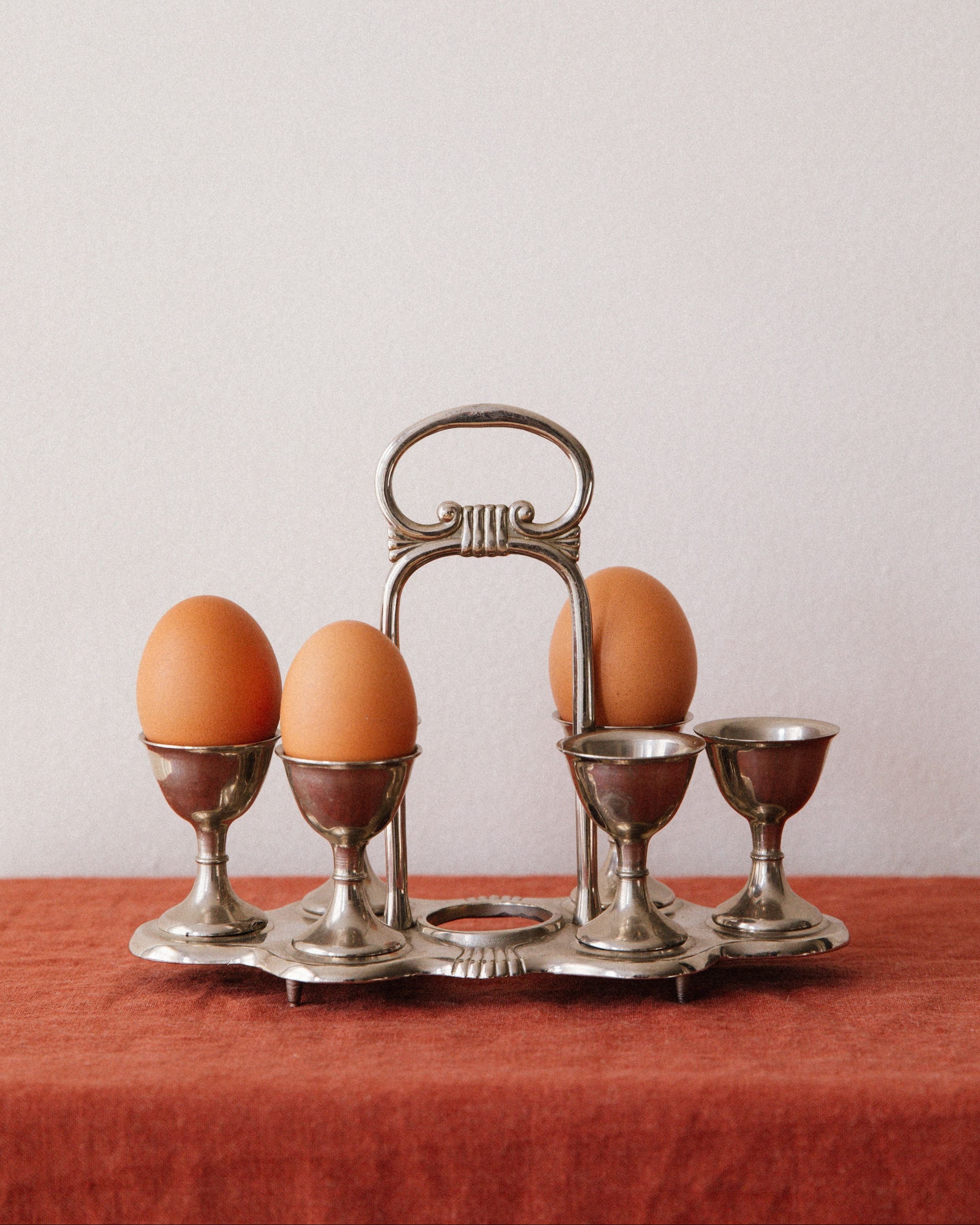 Four brown eggs rest in sculpted cups on the Arcā Collections Vintage Egg Cruet Holder Set, a silver-plated accent atop a rust cloth, creating an elegant tabletop display against a plain light background.