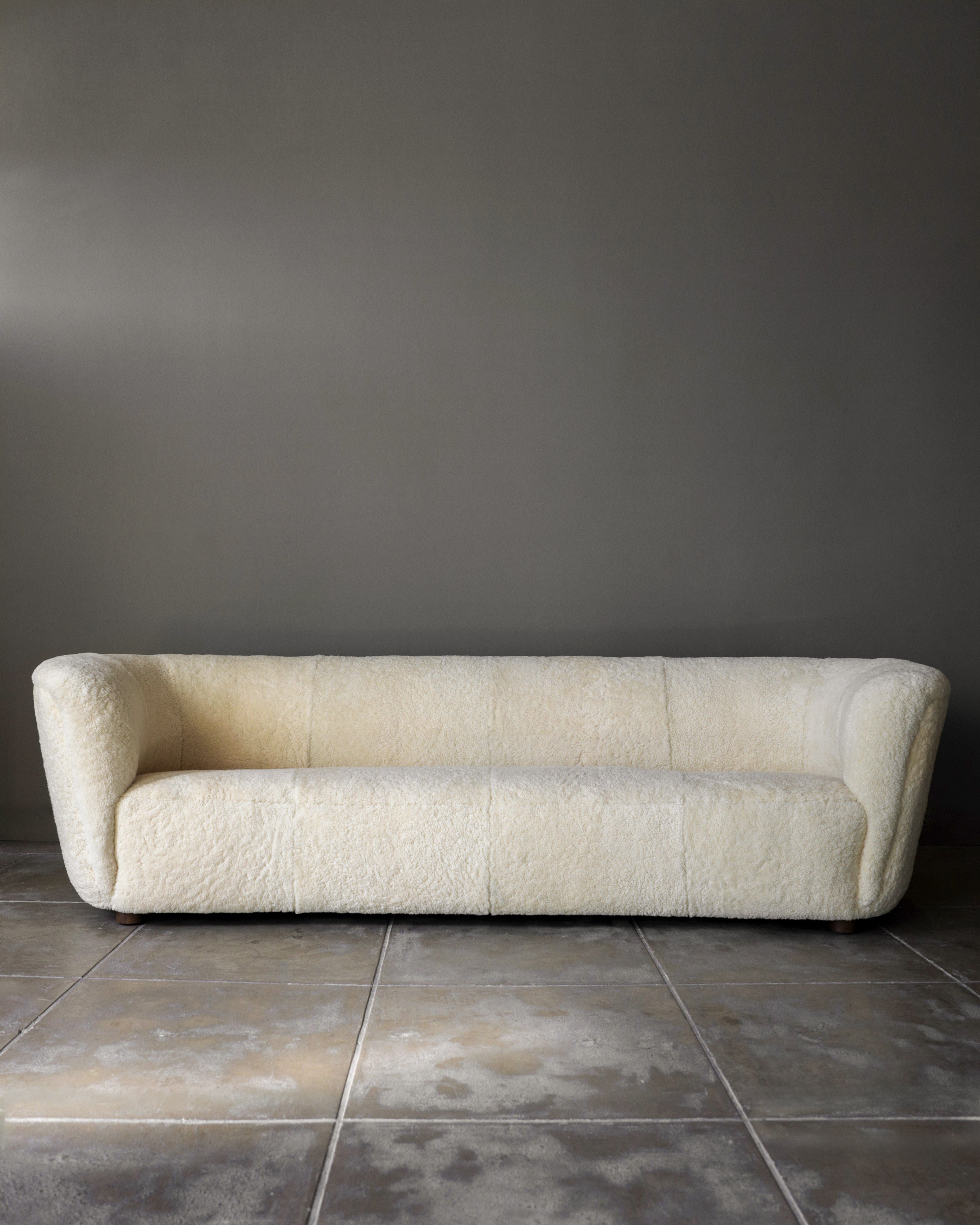 A Fritz Hansen Sofa in Shearling by Studio OSKLO, featuring a cream, textured finish, sits against a plain gray wall on a tiled concrete floor.