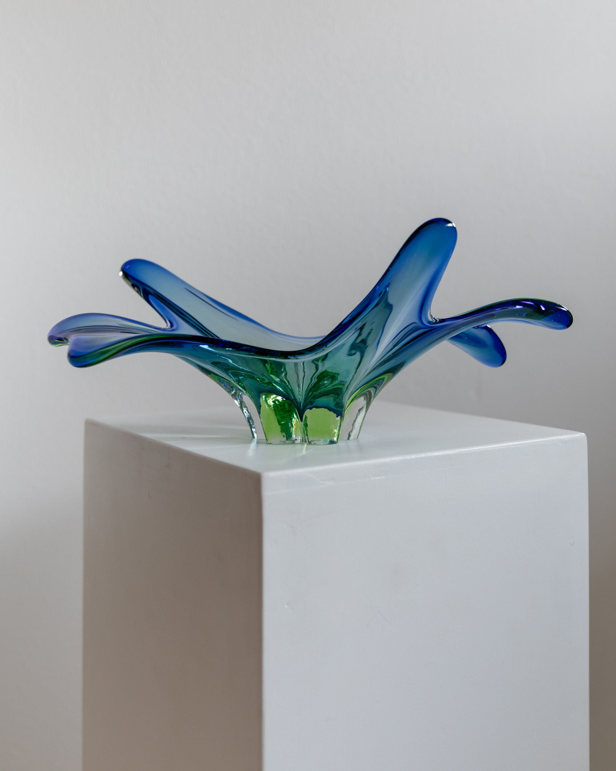The Bottega Jacobs Murano Glass Sommerso Centerpiece 70s, with its stunning blue and green wave-like extensions, sits gracefully on a white pedestal against a plain background. This vintage Murano glass piece is sure to captivate art lovers everywhere.