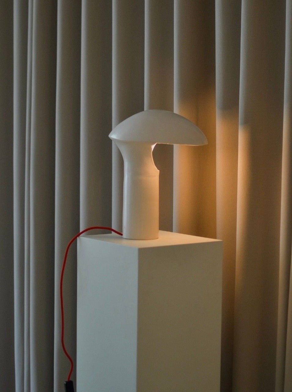 A modern, white mushroom-shaped table lamp with a red power cord sits atop a tall white pedestal. The N11 Lamp by Garado Studios, reminiscent of lamps found in contemporary spaces like the Niemeyer Center, is illuminated against a backdrop of beige curtains, creating a soft, warm atmosphere.