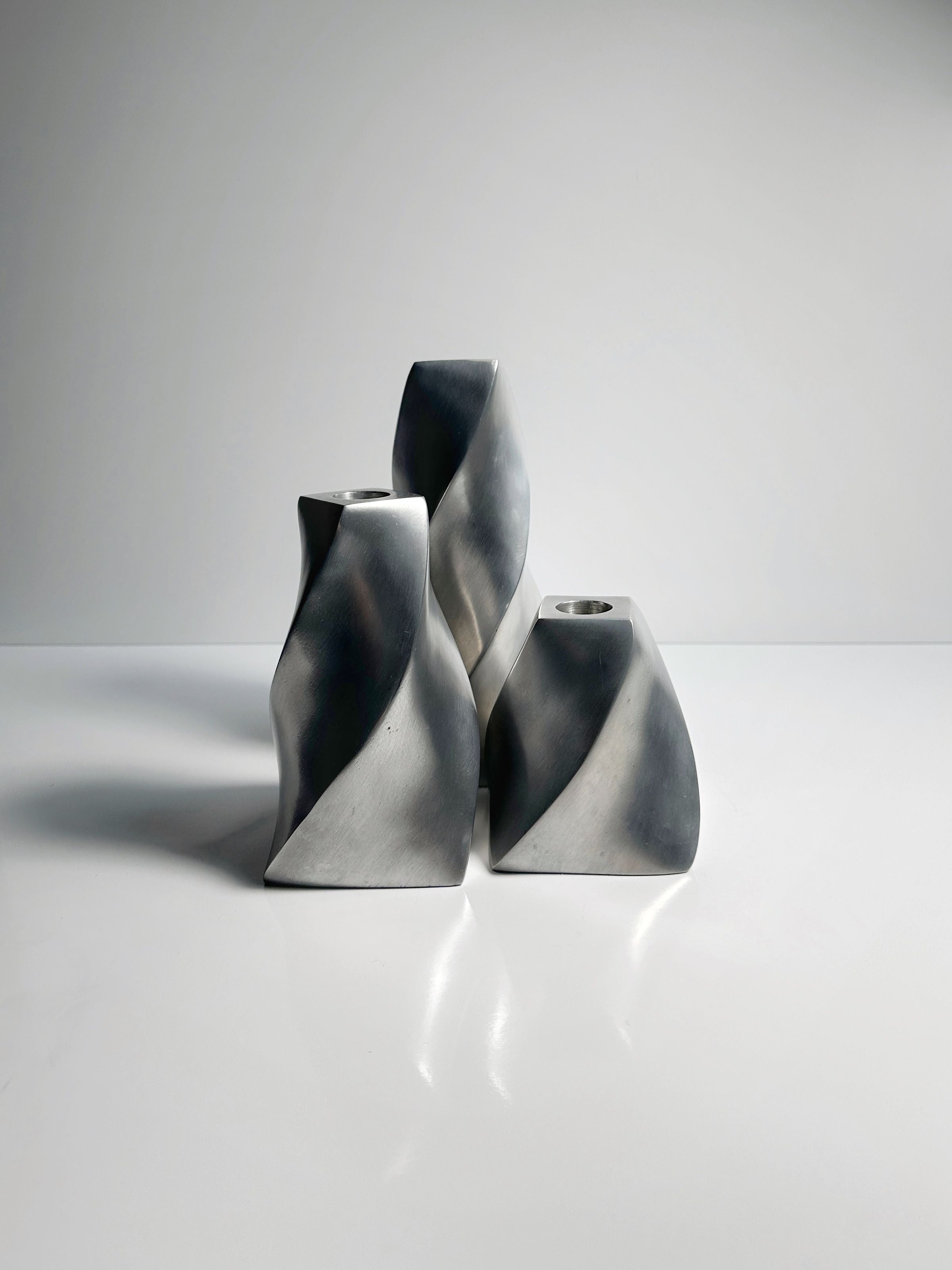 A set of three Dodo Vintage aluminium twisted candle holders rests on a smooth white surface against a plain backdrop. Their spiraled form casts subtle shadows, contributing to a minimalist and contemporary aesthetic.