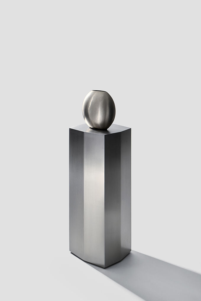 The 'Ame II' Steel Pedestal by Zoé Wolker Studio stands tall and sleek with a metallic, oval-shaped vase atop it, creating an architectural statement against a plain light gray background with a soft right-cast shadow.