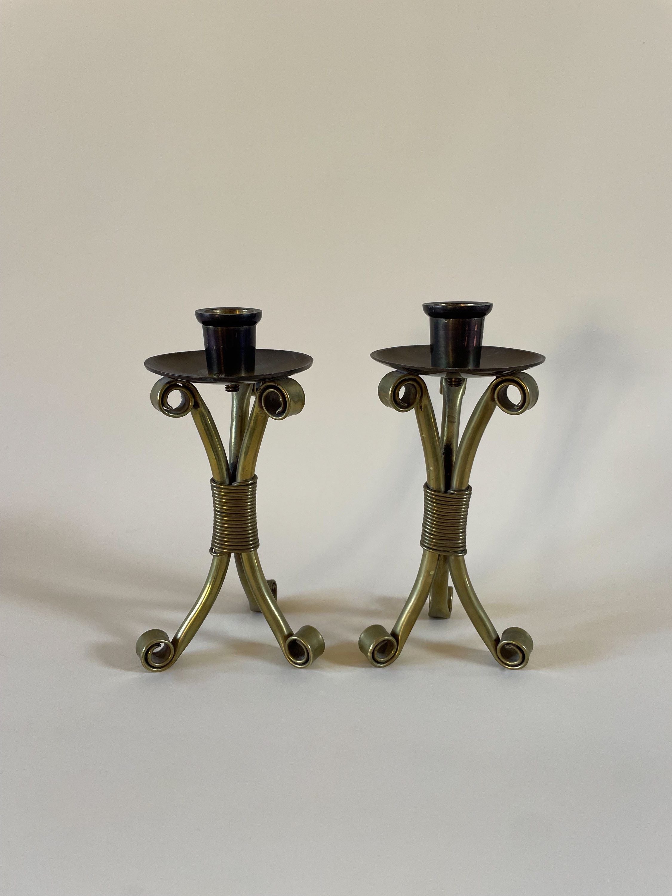 Two antique Danish brass candleholders with intricate, curved designs at the base stand side by side against a plain, light background. The mid-century holders boast a dark, polished finish and sculptural form. These are the **Pair of Mid-Century Danish Candleholders** by **Maud Vaughan**.