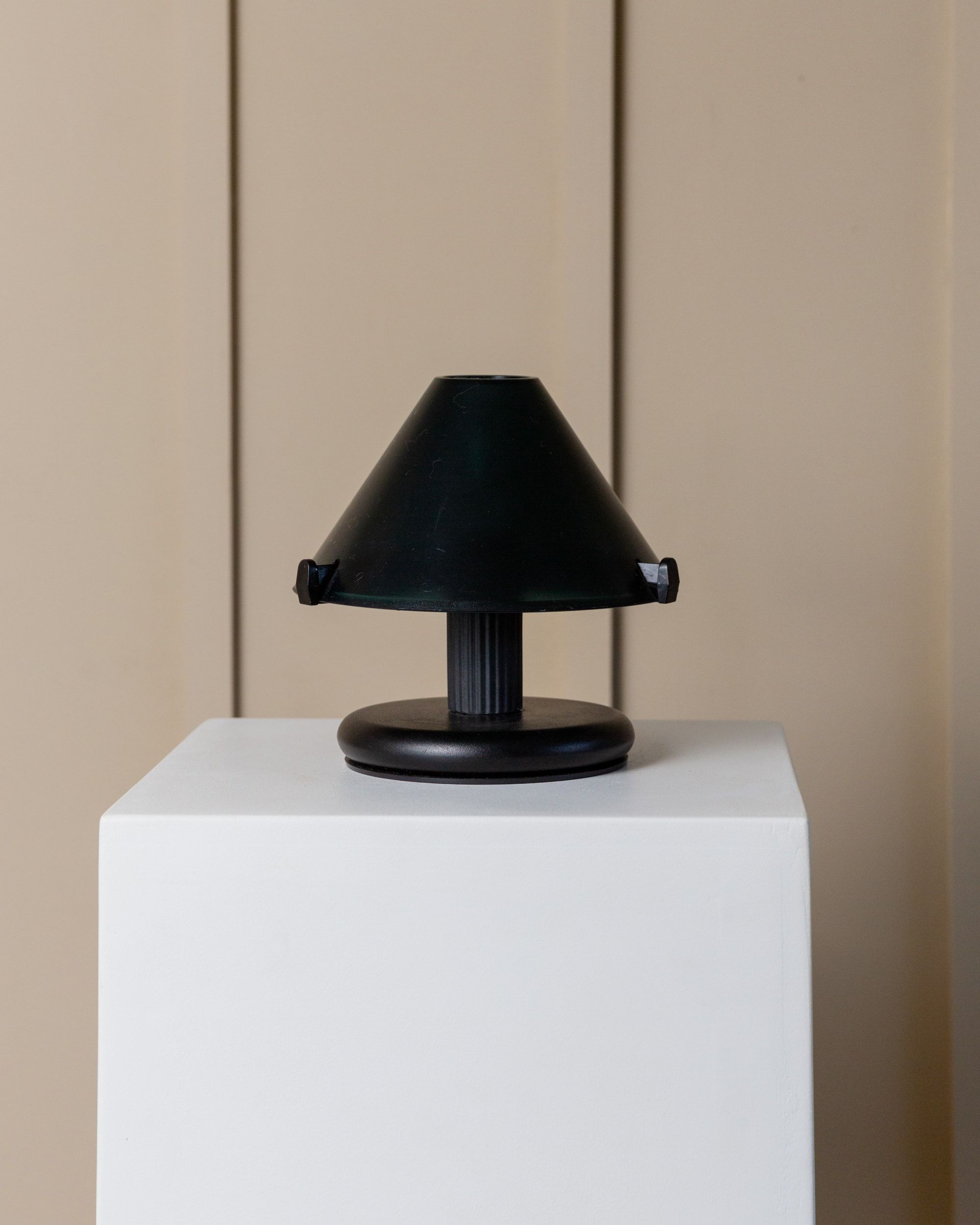 The Vintage Table Lamp 80s by Bottega Jacobs, featuring a small black design with a conical shade, is placed on a white pedestal against a neutral beige background. Its circular base and short ribbed stem emit an air of timeless elegance that enhances any decor.