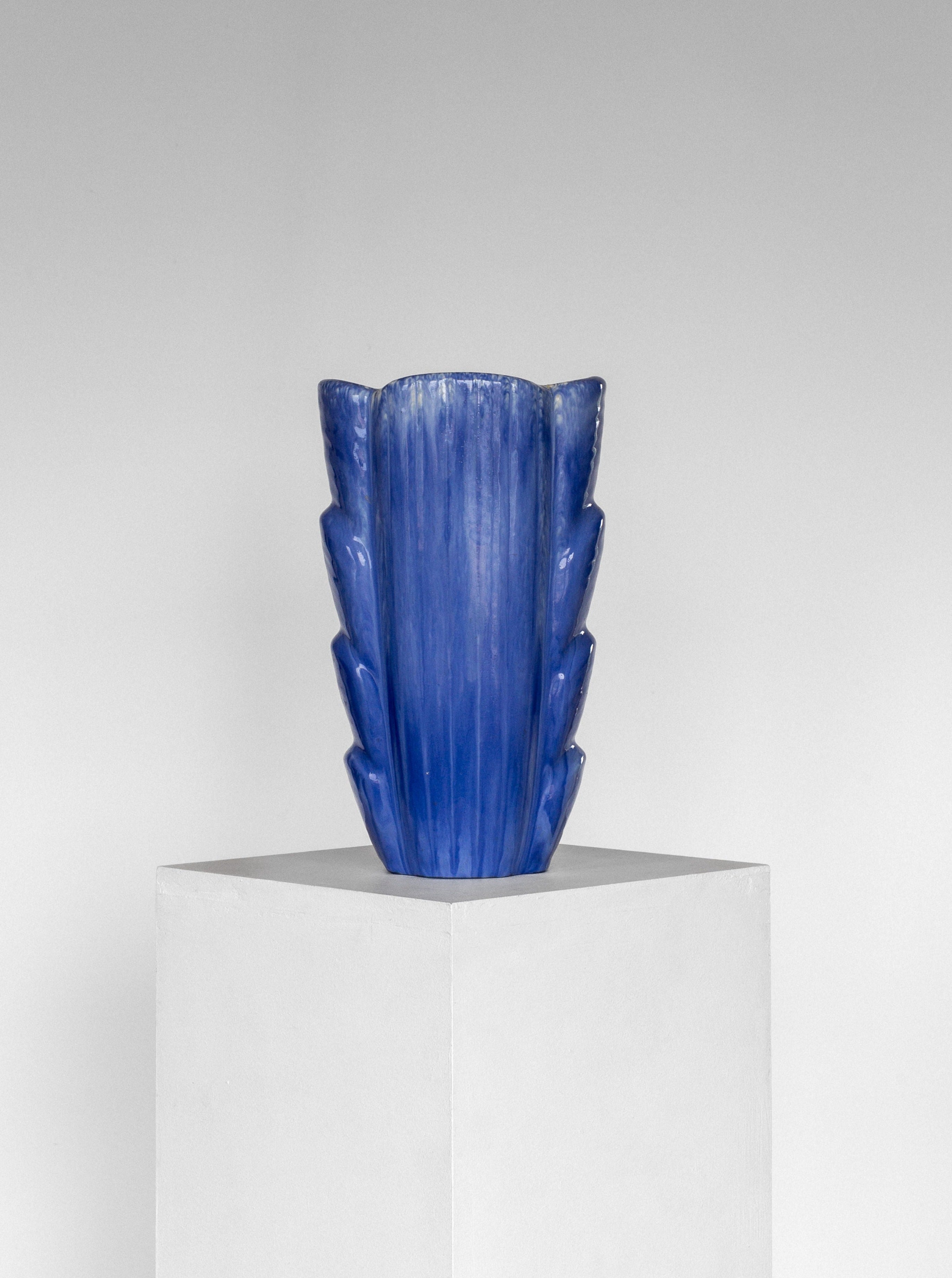 A blue ceramic large vase by Gunnar Nylund, featuring an abstract, geometric design with flared sides that resemble stylized leaves, is showcased on a white cubic pedestal against a plain gray background. This distinctive Spigel piece from the 1960s evokes the charm of Mid-Century Modern ceramic vases found in Scandinavian studios.