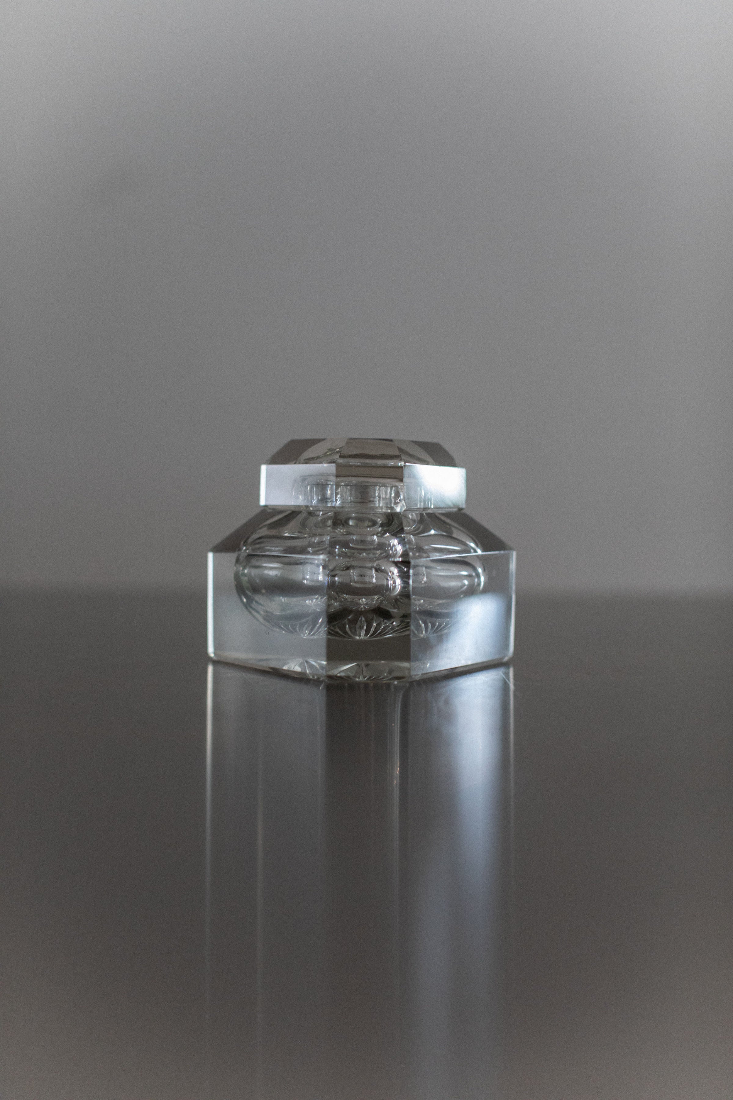 A product from Out For Lunch, the Glass Perfume Bottle features a transparent hexagonal design reminiscent of vintage elegance, artfully displayed on a reflective surface against a gray background. Its geometric form highlights exquisite craftsmanship with elegant reflections and refractions.