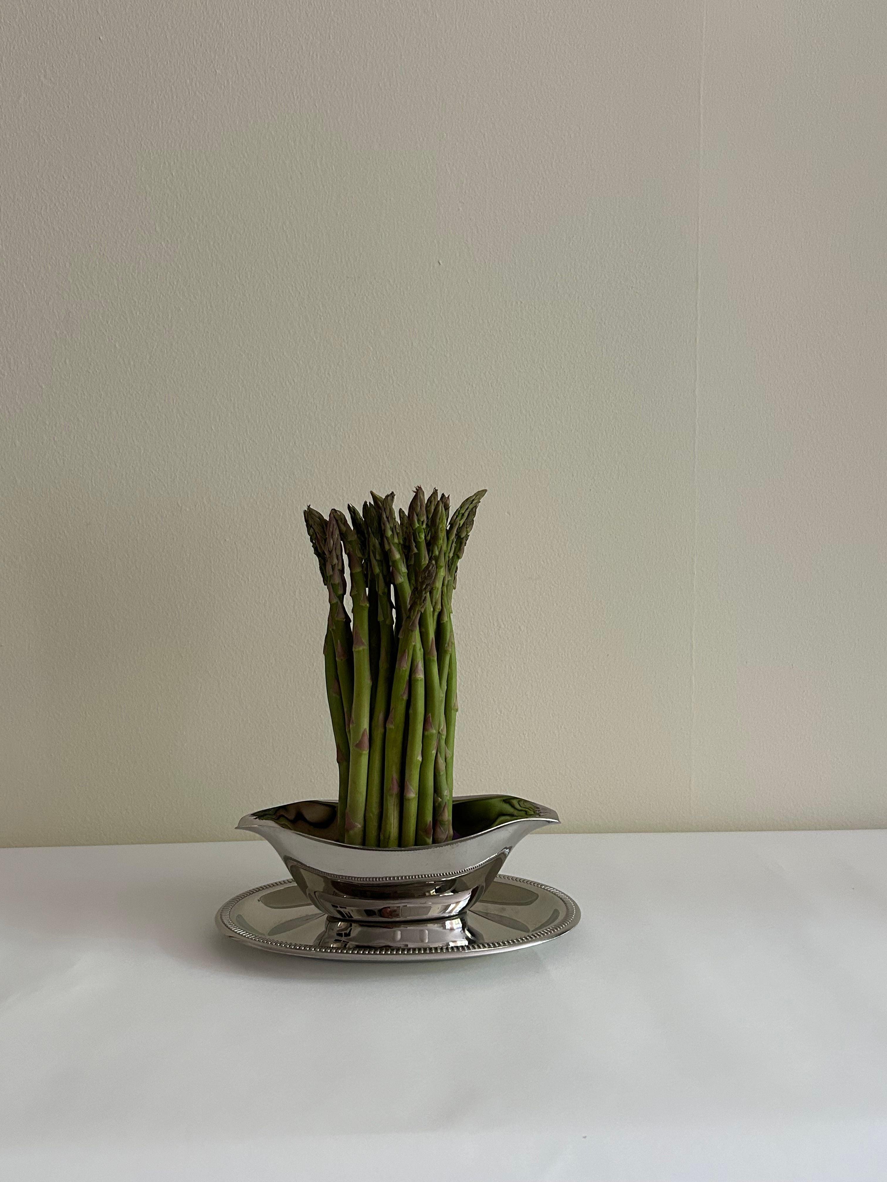 A collection of fresh asparagus spears is elegantly arranged upright in a decorative Flock Studio Stainless Steel Server Sauce dish with a polished finish, resting on a coordinating silver platter against a simple, light-colored background.