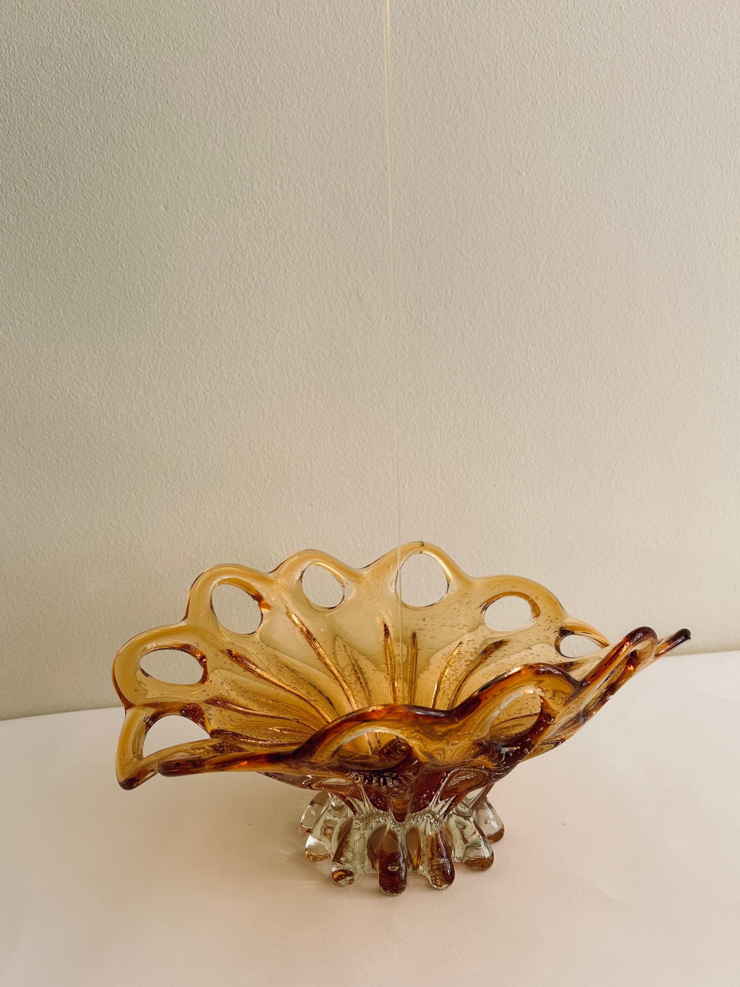 A Murano Glass Amber Bowl by Flock Studio, featuring a wavy, scalloped edge and intricate cut-out designs, handcrafted with Italian glass artistry. It is displayed on a white surface against a light-colored background.