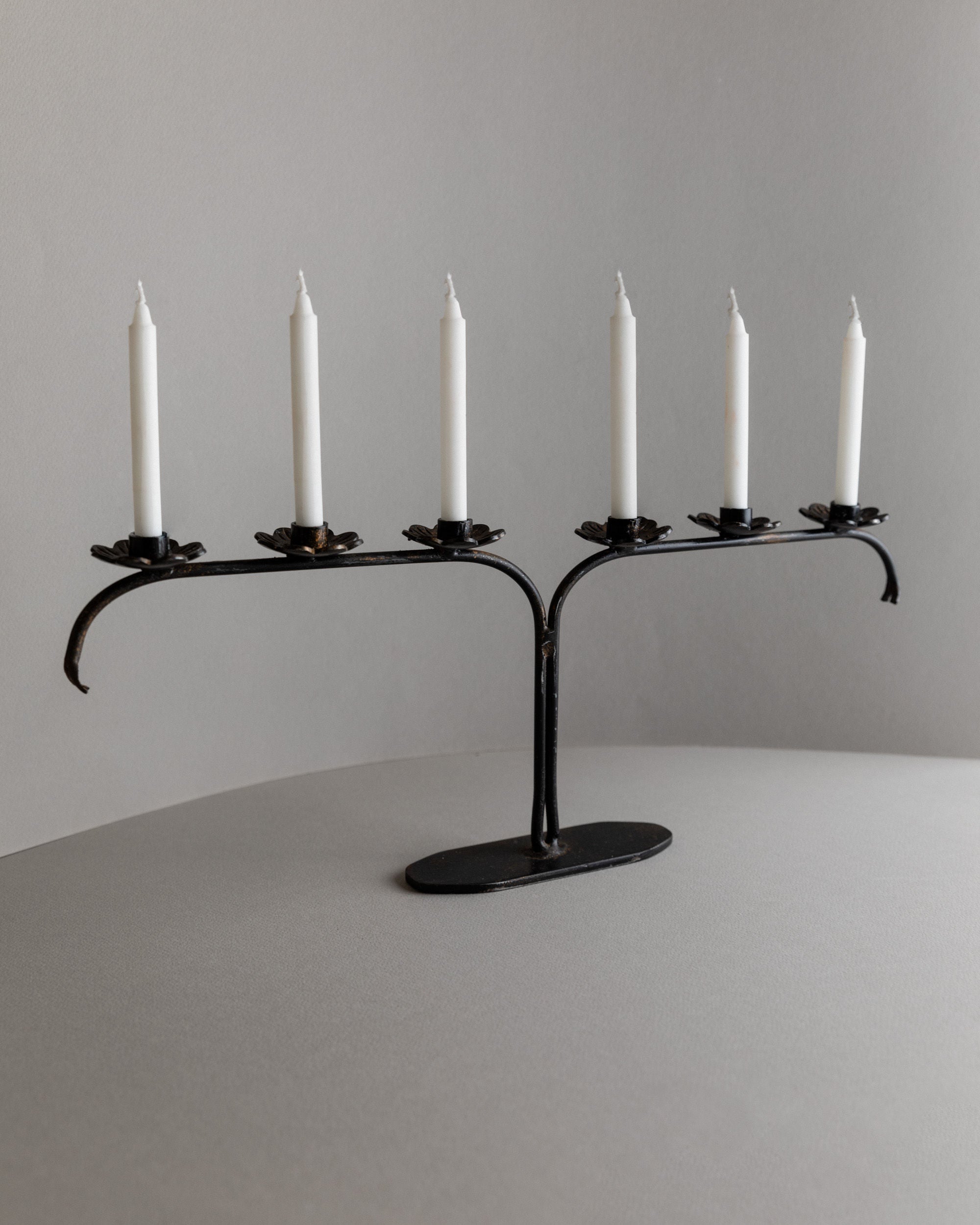 The Iron Candleholder from Bottega Jacobs, Italy 1960s, is a black metal piece with an L-shaped design that elegantly holds five unlit white taper candles. Its minimalist brutalist style complements any neutral gray surface against a gray background.