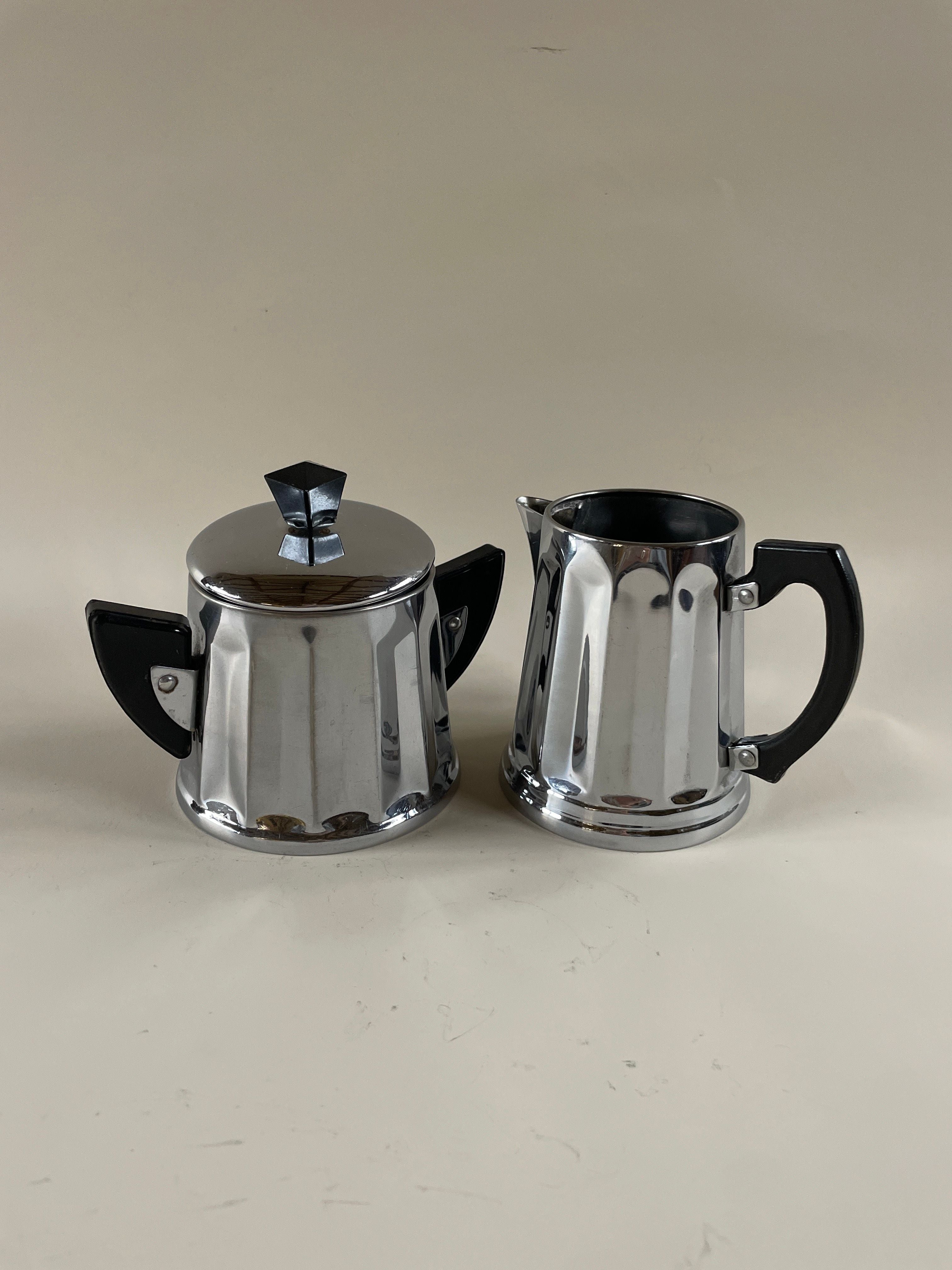 A retro Art Deco teapot and sugar bowl set by Maud Vaughan, adorned with sleek Bakerlite handles and a geometric design set against a simple beige backdrop.