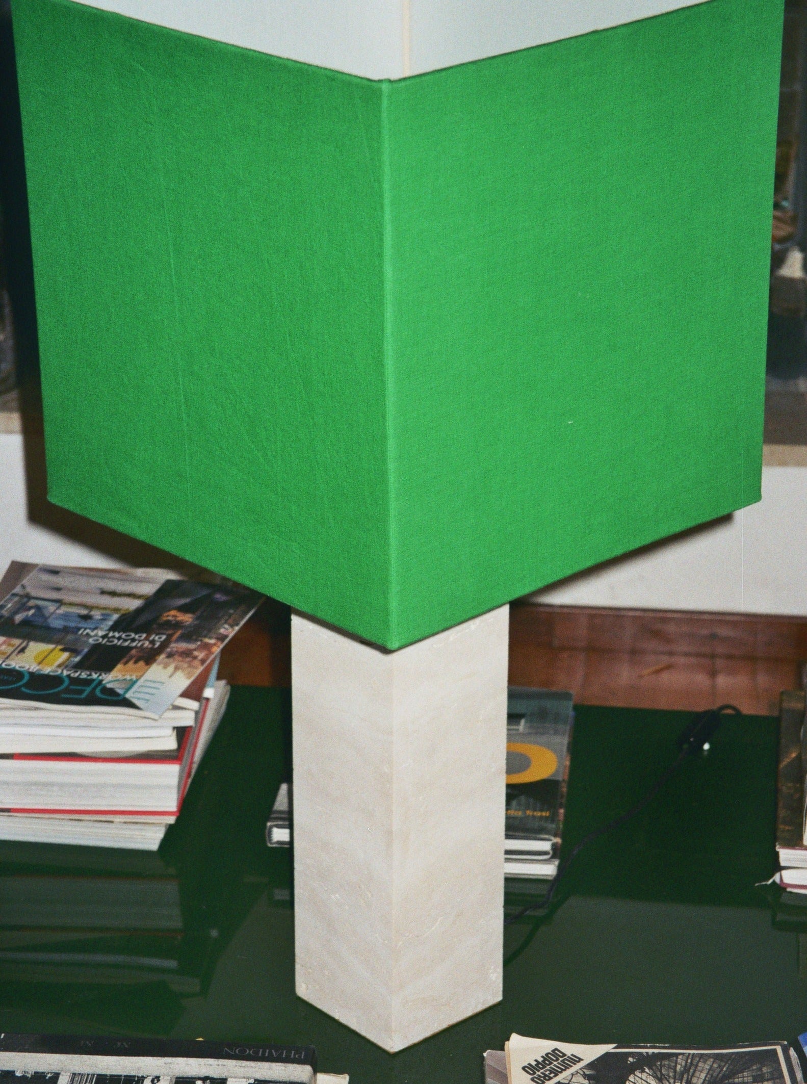 A Roma I Lamp Squared White Travertine with a green linen shade, a limited edition by Nassi Lamps, sits on a glass surface. Surrounding the lamp are numerous books and magazines stacked in various piles, creating an inviting reading nook.