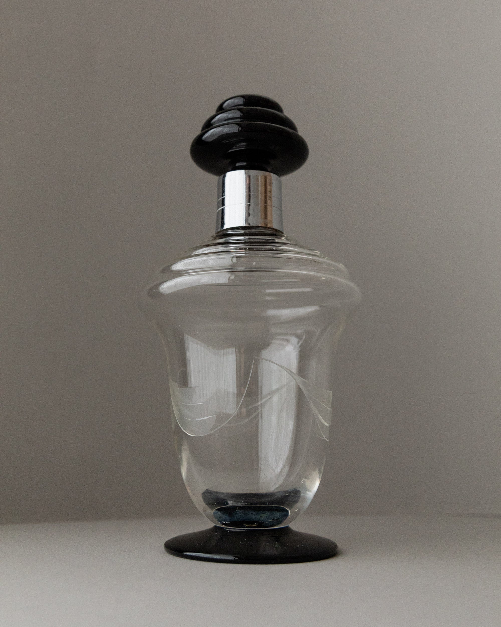 A Bottega Jacobs Murano Glass Decanter from the 1930s, featuring etched swirl patterns and a decorative black lid, sits on a neutral surface, reminiscent of vintage elegance.