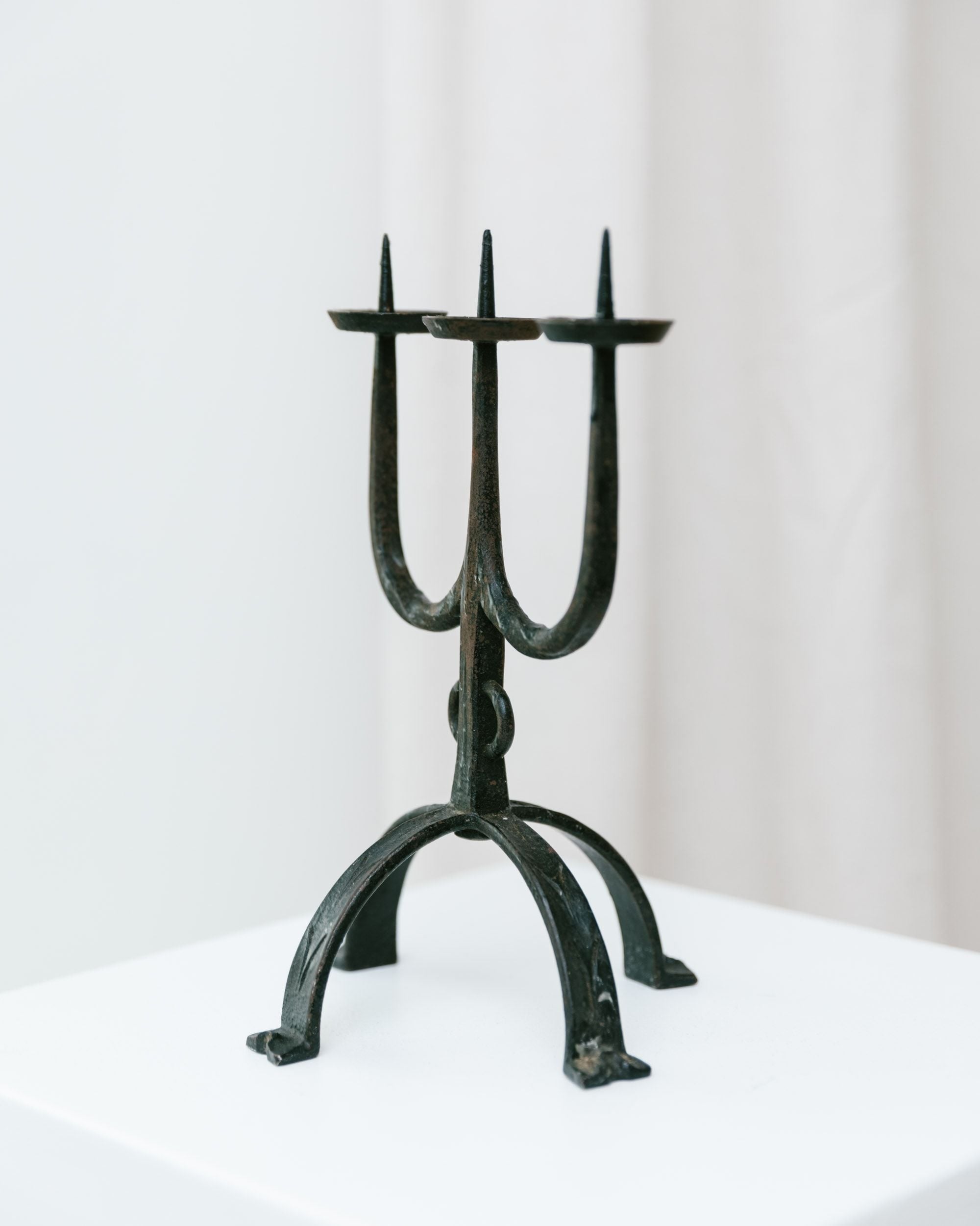 The Iron Candleholder 70s by Bottega Jacobs is elegantly showcased on a white pedestal. This vintage sculptural piece boasts three upward-pointing prongs with candle holders and a tripod base, all set against a plain white background, exuding an air of elegance and timeless charm.
