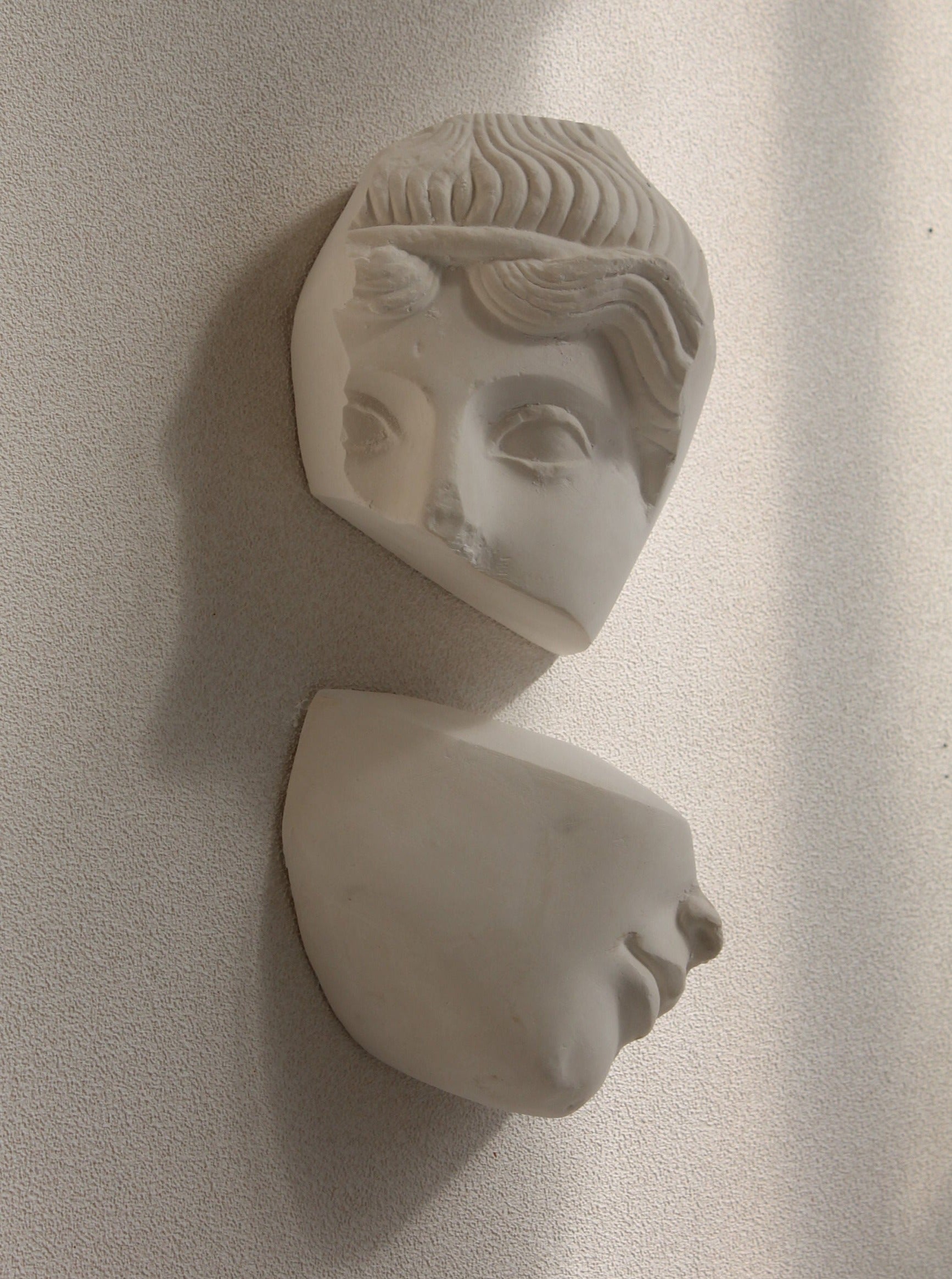 A photo of the Diptyque Géométrique Sculpture by Marina Mankarios, mounted on a textured beige wall. Expertly crafted, the white marble statue's head is divided into two pieces: the top portion showcasing the eyes and forehead, and the bottom portion displaying the nose and mouth, with visible cracks revealing its white patina plaster.