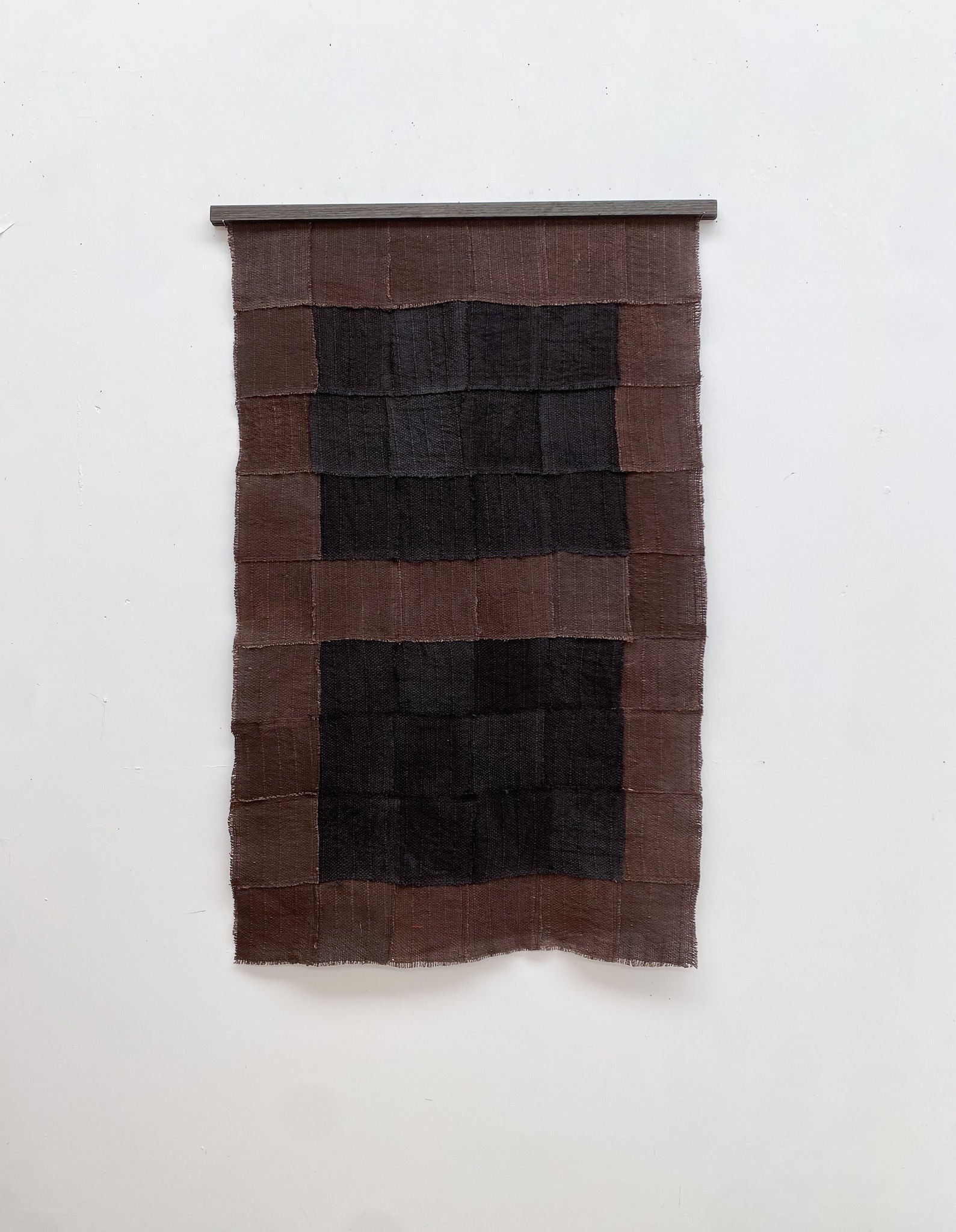 The Ground Echo textile wall hanging by Sundance Studio, inspired by sweeping plains, features a grid of black and brown rectangles. Its woven fabric texture hangs from a horizontal rod against a white backdrop, blending art and nature.